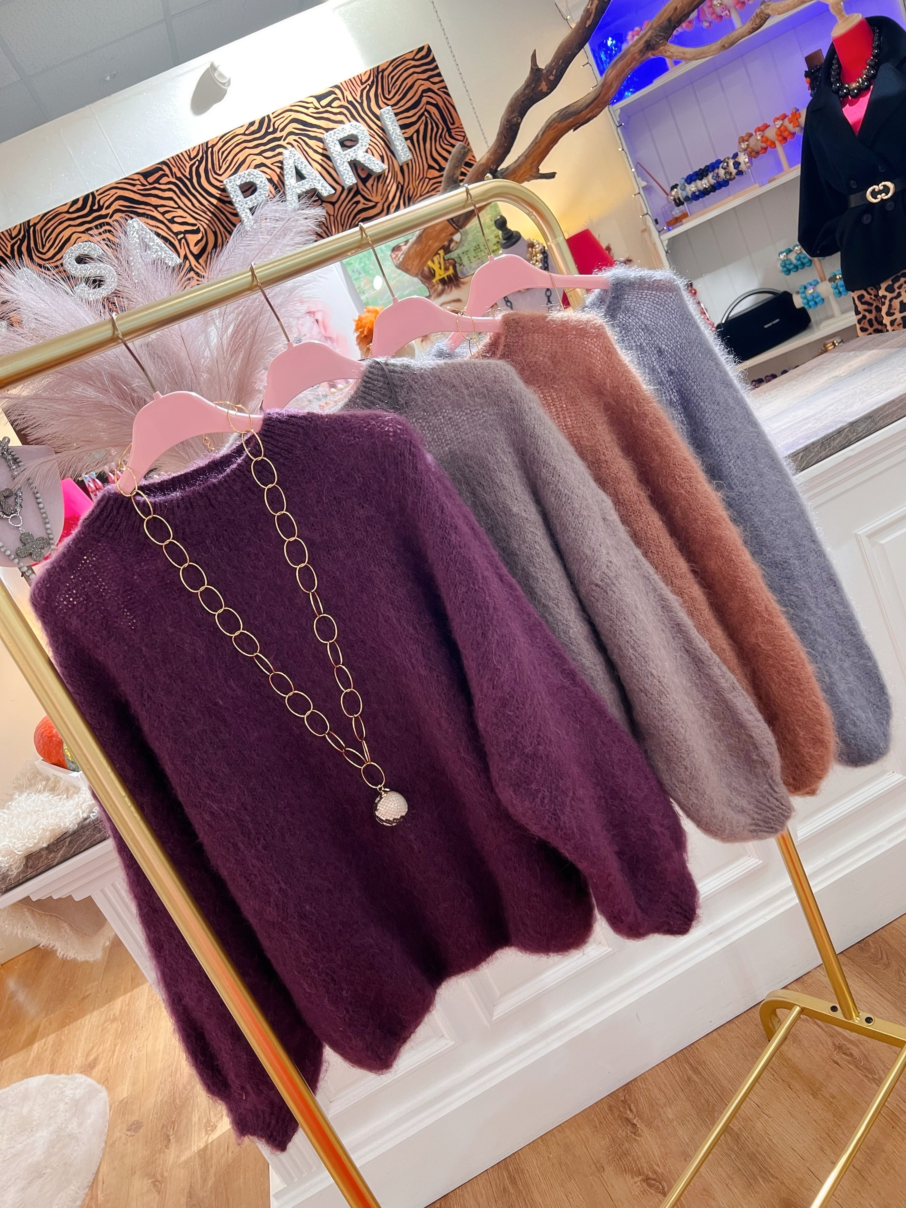 Vier weiche Pullover aus Kid Mohair in warmen Herbsts Tönen. Violet mit Kette, Taupe, Karamell und Grau. Hängen auf roséfarbenen Kleiderbügel an einer goldenen Stange in einer Boutique.