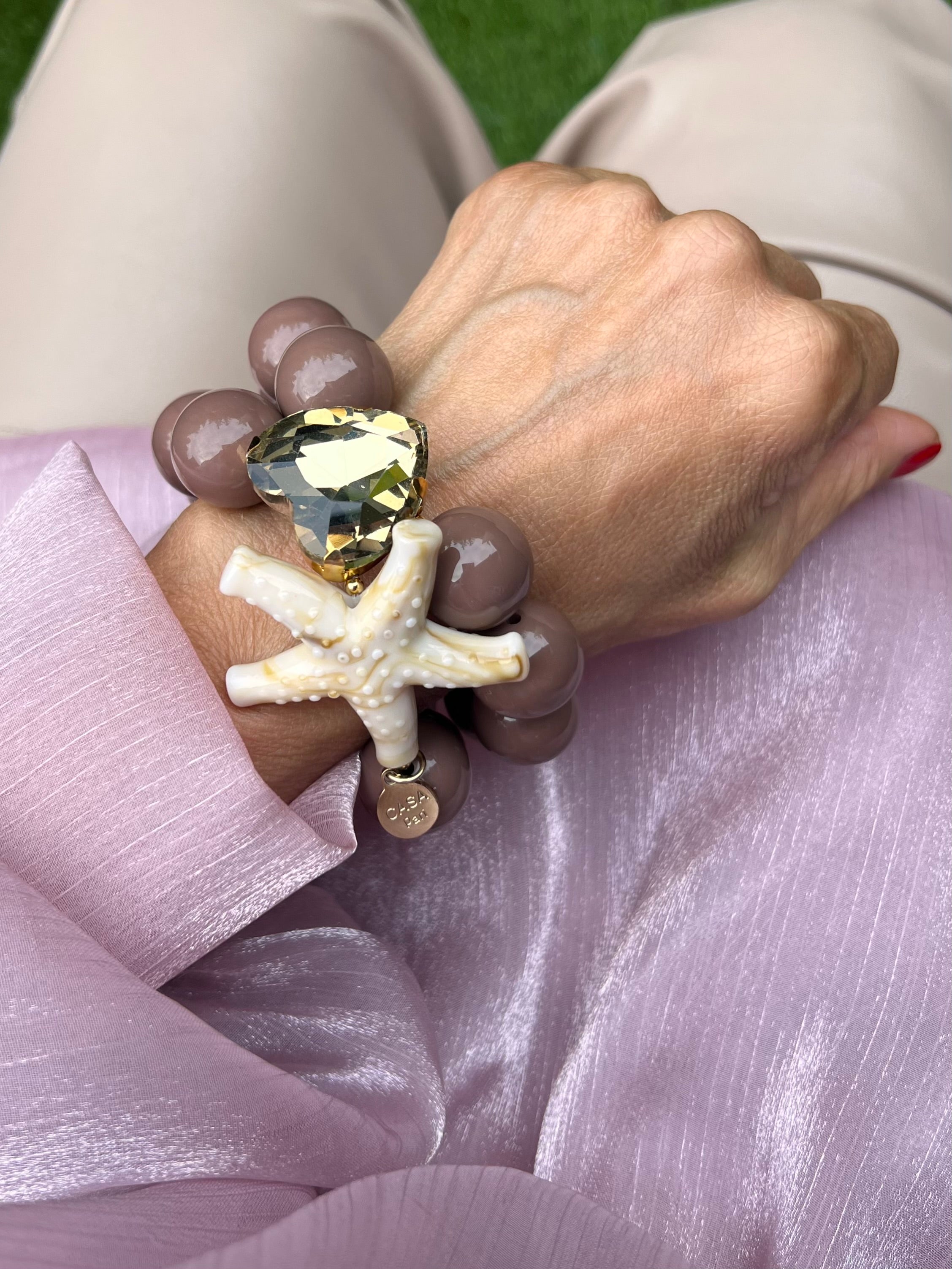 Armband mit zwei Reihen glänzender Perlen in Taupe, verziert mit einem funkelndem Kristalle Herz in Champagner, auf hellem Hintergrund.