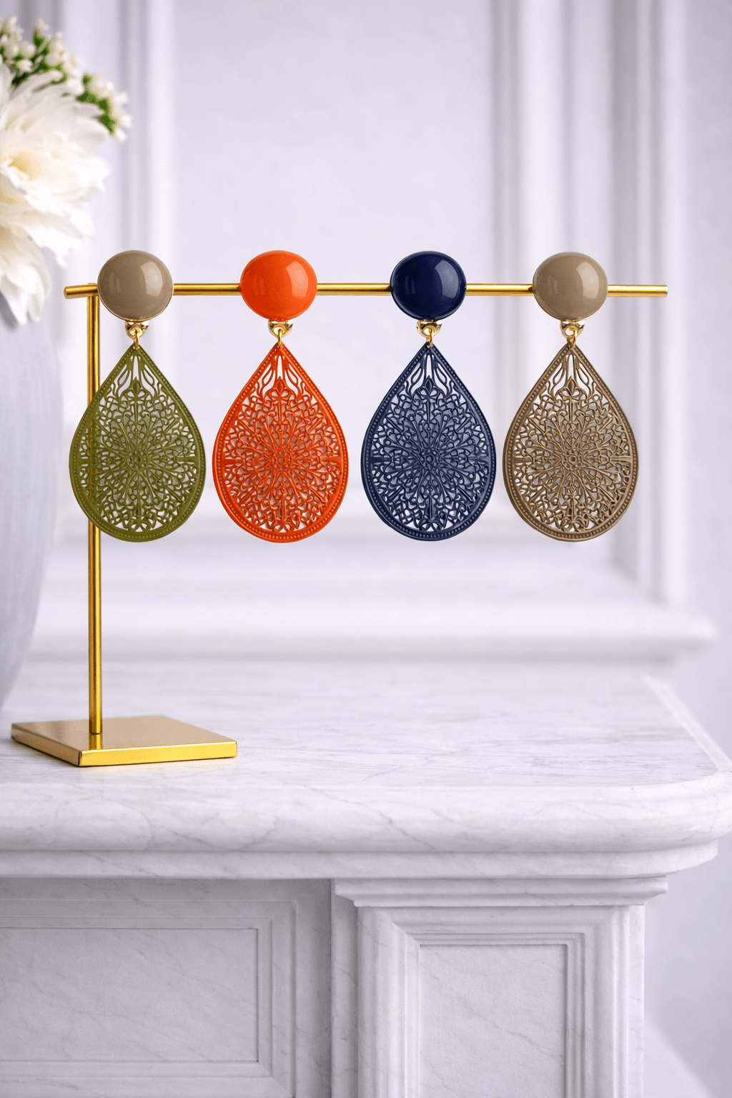 Clip-Ohrringe mit rundem, glänzendem Stecker und tropfenförmigem Anhänger aus fein verziertem Metall, in Olivgrün, Orange, Dunkelblau und Taupe, präsentiert auf goldenem Schmuckständer vor hellem Hintergrund.