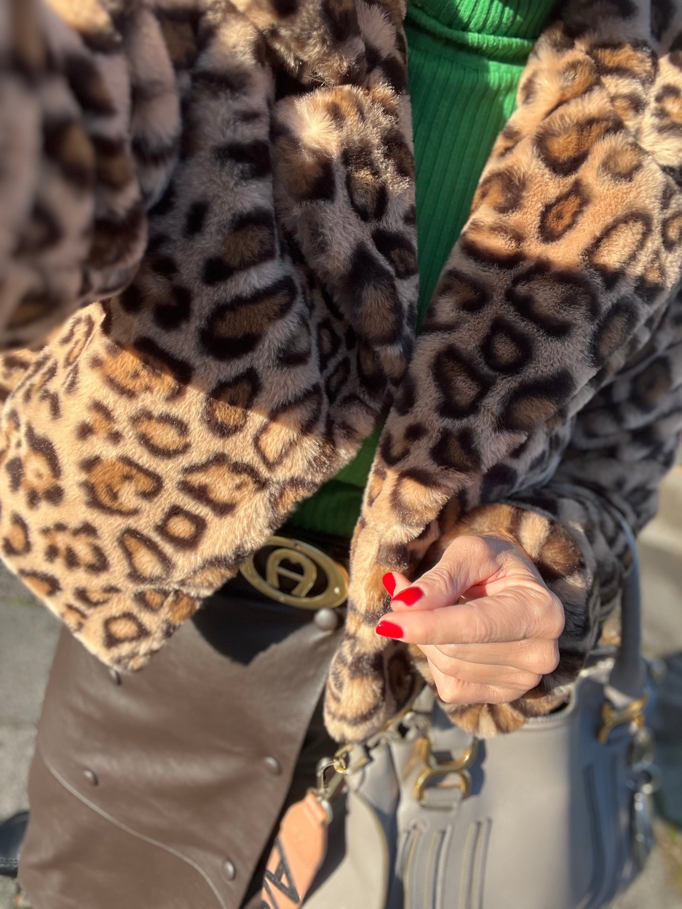 Nahaufnahme eines Outfits. Zu sehen ist eine Jacke mit Leopardenmuster, ein grüner Strickpullover, ein brauner rock, ein goldfarbener Gürtel und eine graue Handtasche. Die Hand der Person ragt ins Bild und hat rote, lackierte Fingernägel.