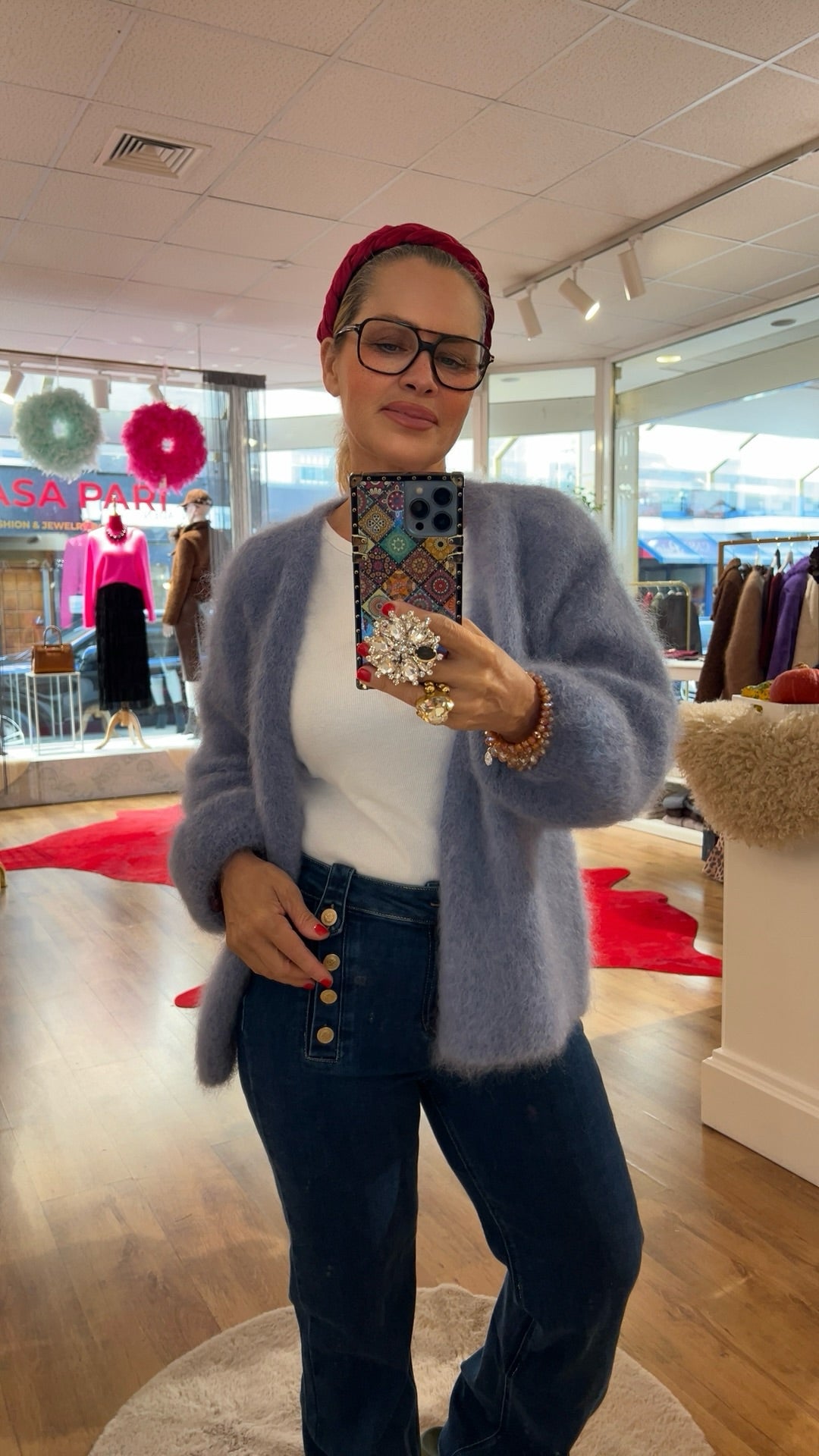 Frau trägt flauschige Strickjacke in Taubenblau, kombiniert mit weißem Shirt und dunkelblauer Jeans mit Knopfleiste. Im Hintergrund bunte Mode und rote Akzente.