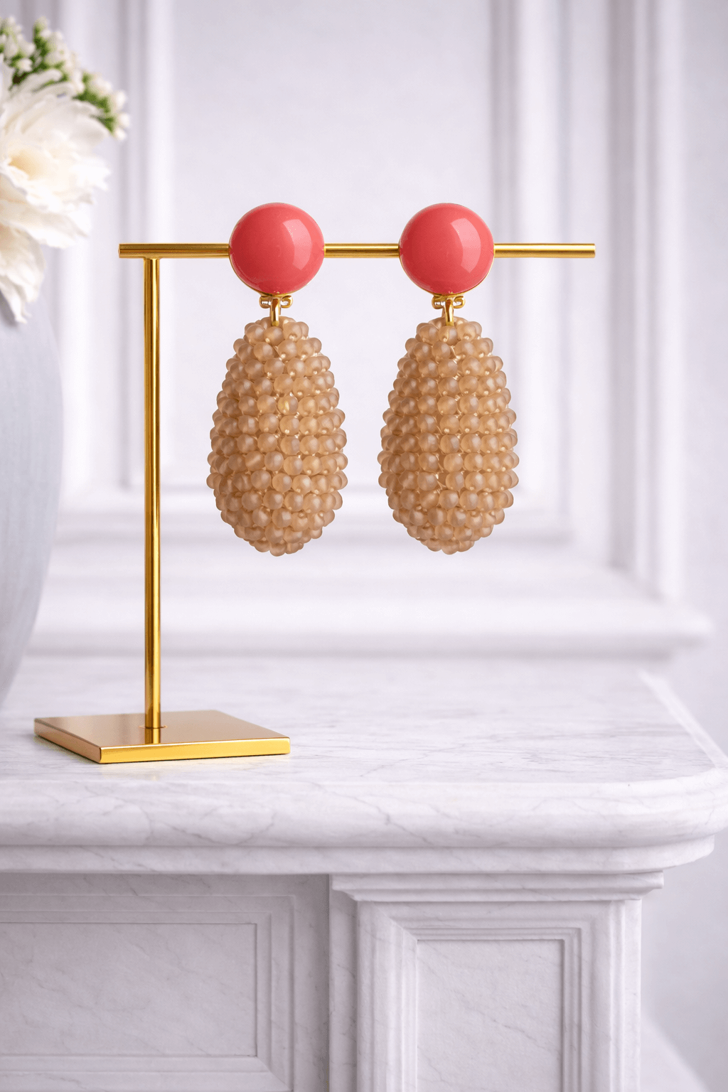 Paar tropfenförmige Ohrringe mit korallfarbenem runden Stecker und beigefarbenem, strukturierendem Perlenkörper auf goldenem Schmuckständer vor hellem Hintergrund.

