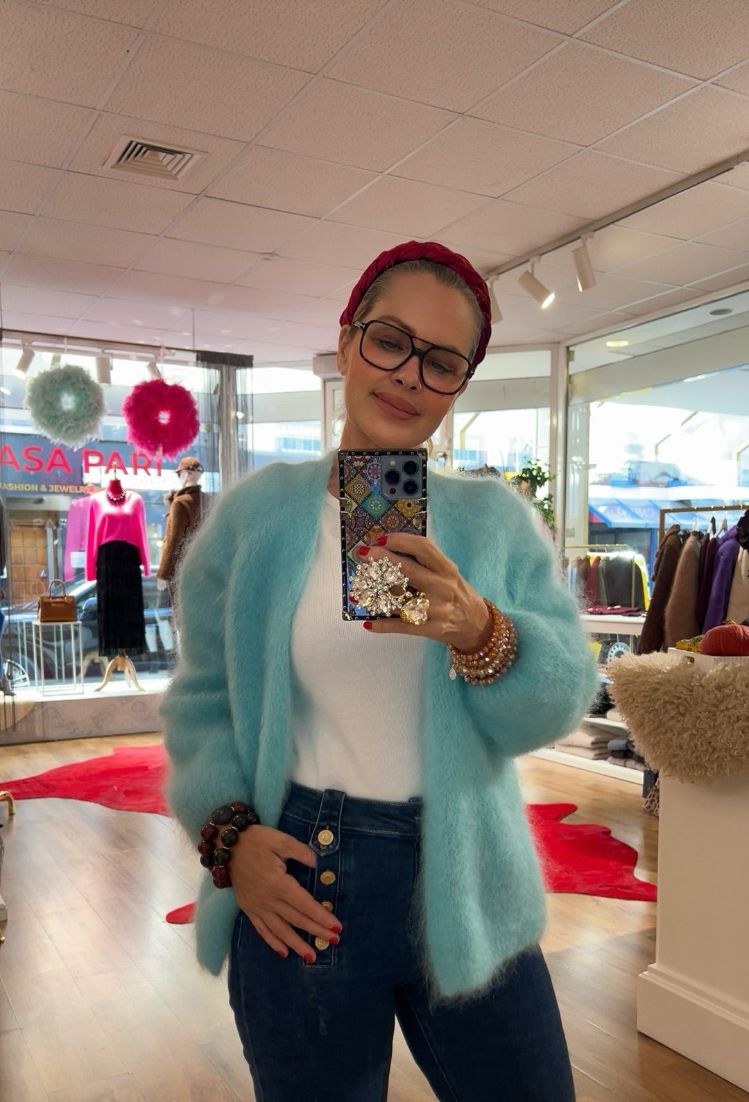 Frau trägt flauschige Strickjacke in Aqua, kombiniert mit weißem Shirt und dunkelblauer Jeans mit Knopfleiste. Im Hintergrund bunte Mode und rote Akzente.