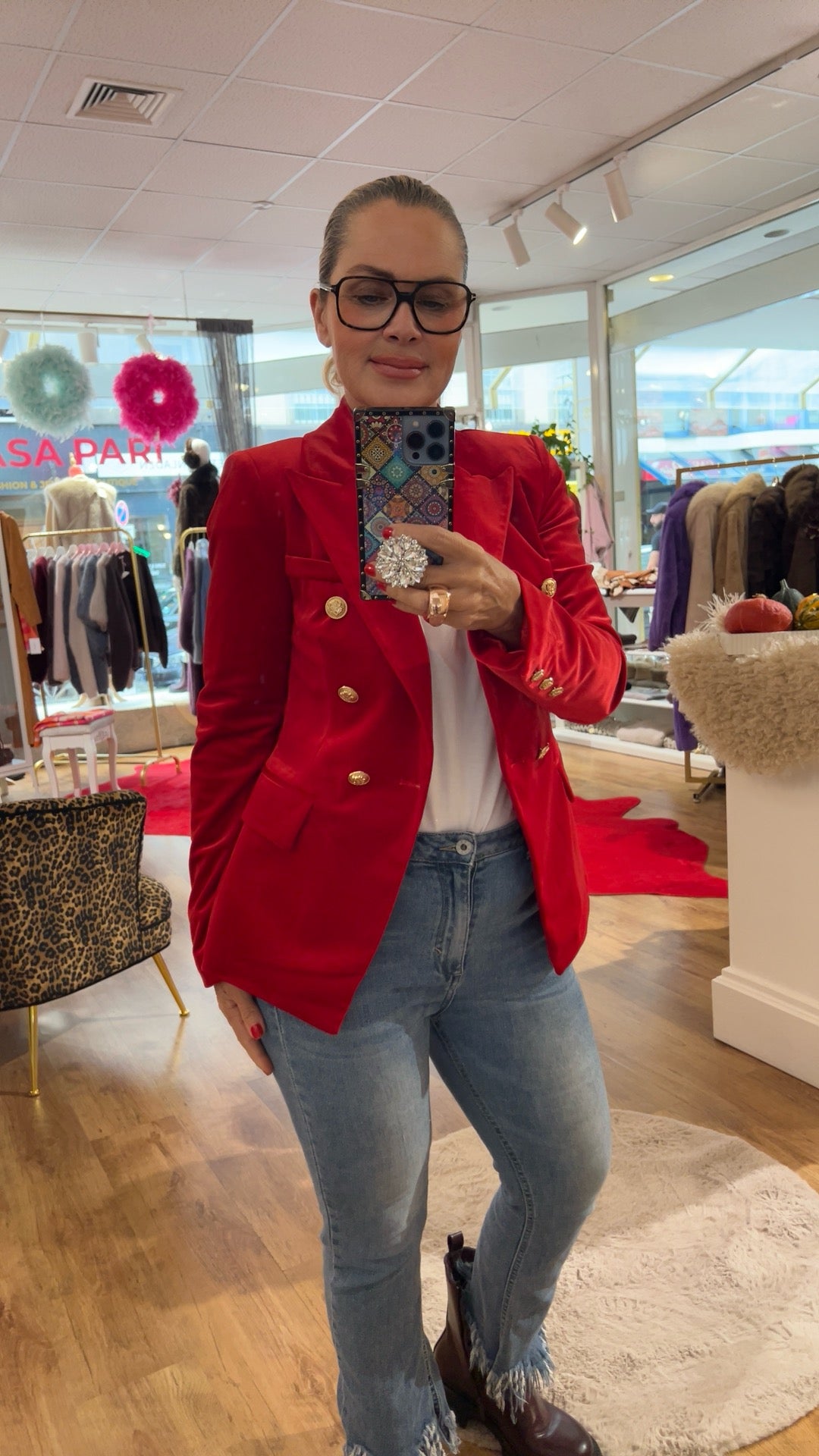 Frau trägt roten Samtblazer mit goldenen Knöpfen. Dazu eine Jeans mit braunen Boots. Sie hält dein Handy in der Hand. Fotografiert in einer Boutique.