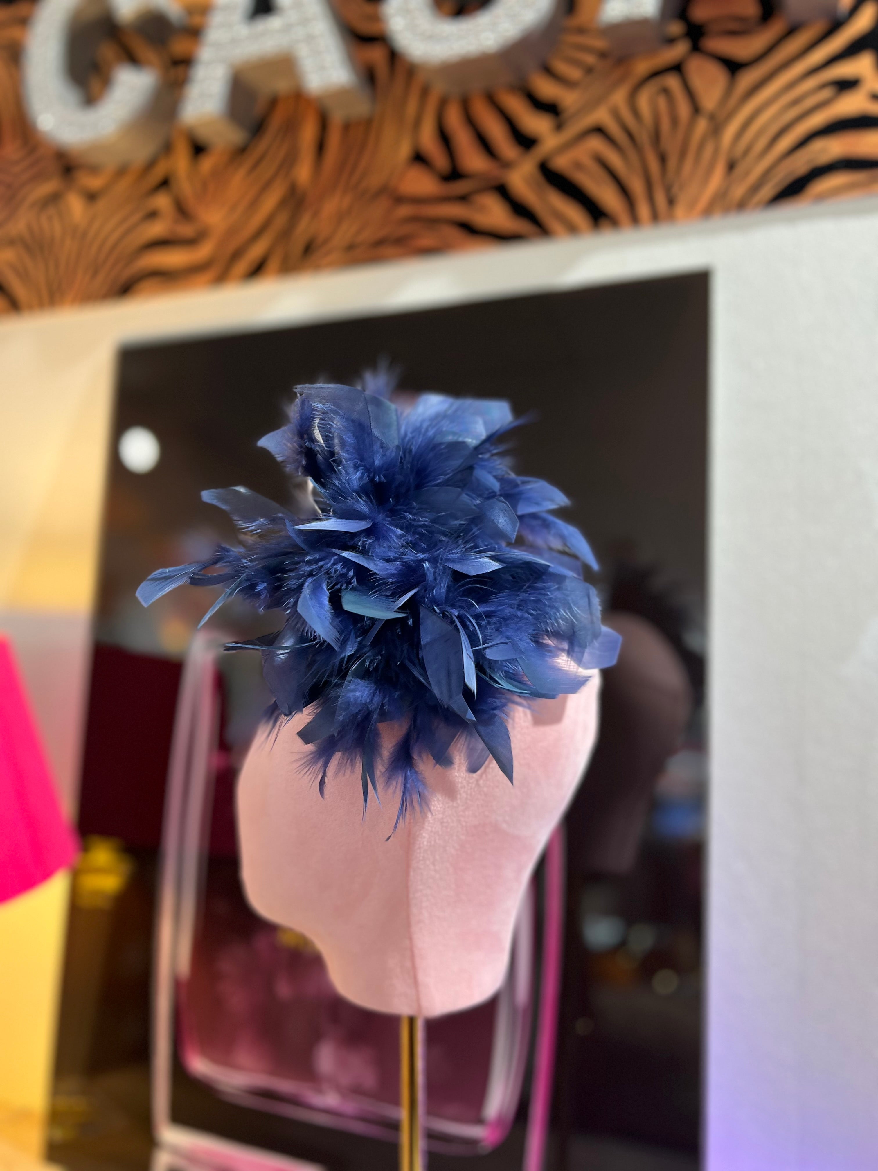 Federbrosche mit großer Blüte aus blauen Federn, befestigt auf einer rosa Stoffpuppe vor dekorativen Hintergrund mit Parfumflakon Motiv.