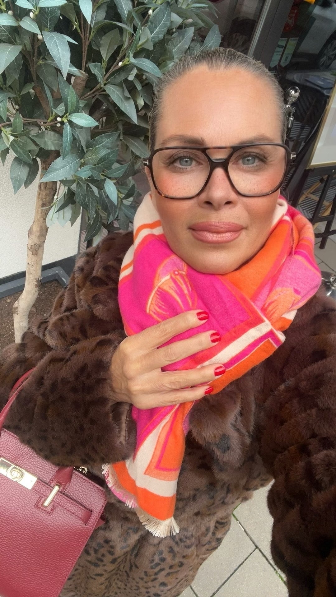 Frau trägt Leo Kunst Fellmantel in Braun, mit roter Tasche und pink, orange, beigem Schal. Und große Brille. Im Hintergrund grüne Pflanzen.
