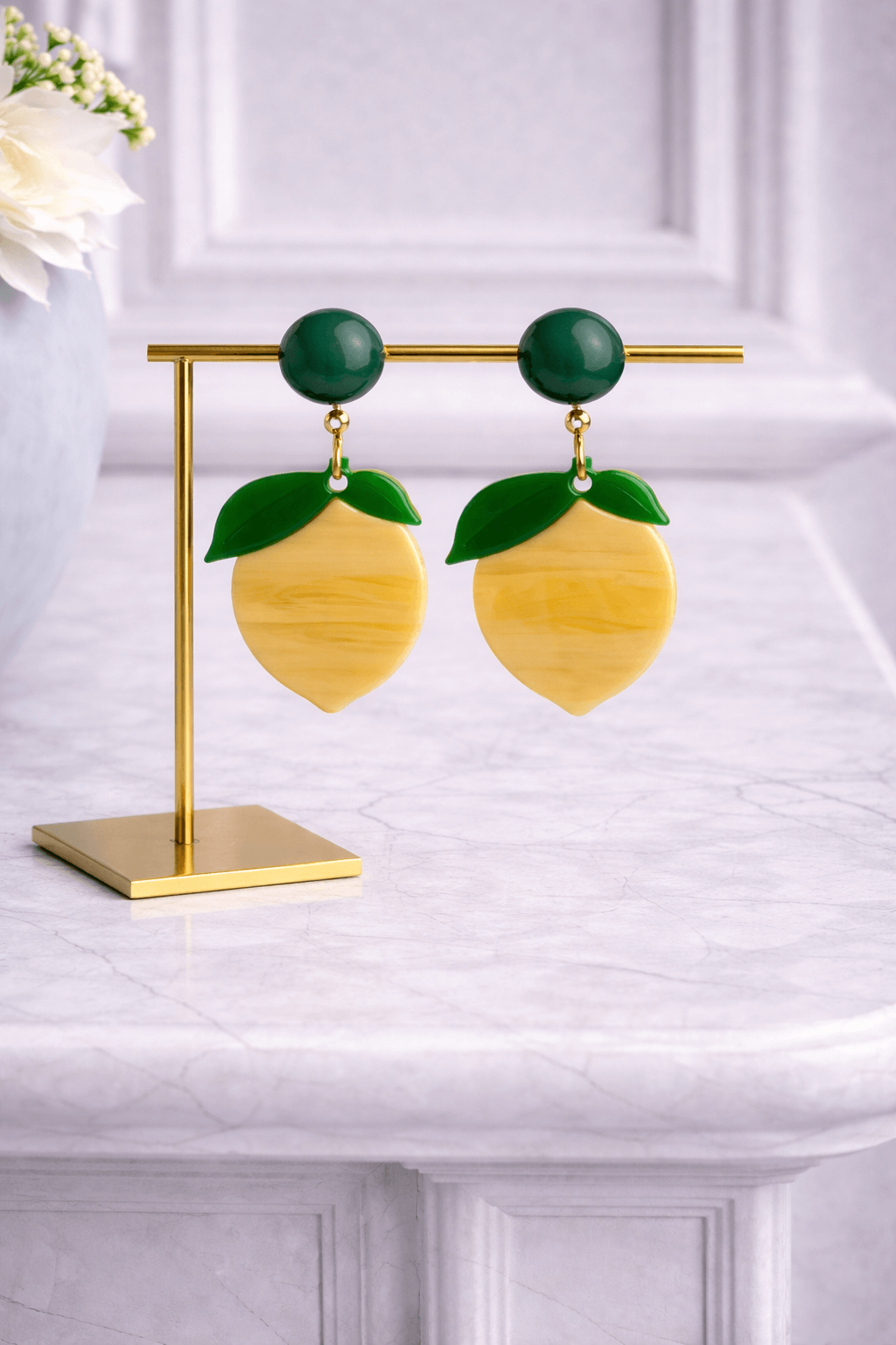 Gelbe Clip-Ohrringe in Zitronenform mit grünem Blatt und rundem, dunkelgrünem Stecker, hängend an einem goldfarbenen Schmuckständer auf einer hellen Marmoroberfläche.