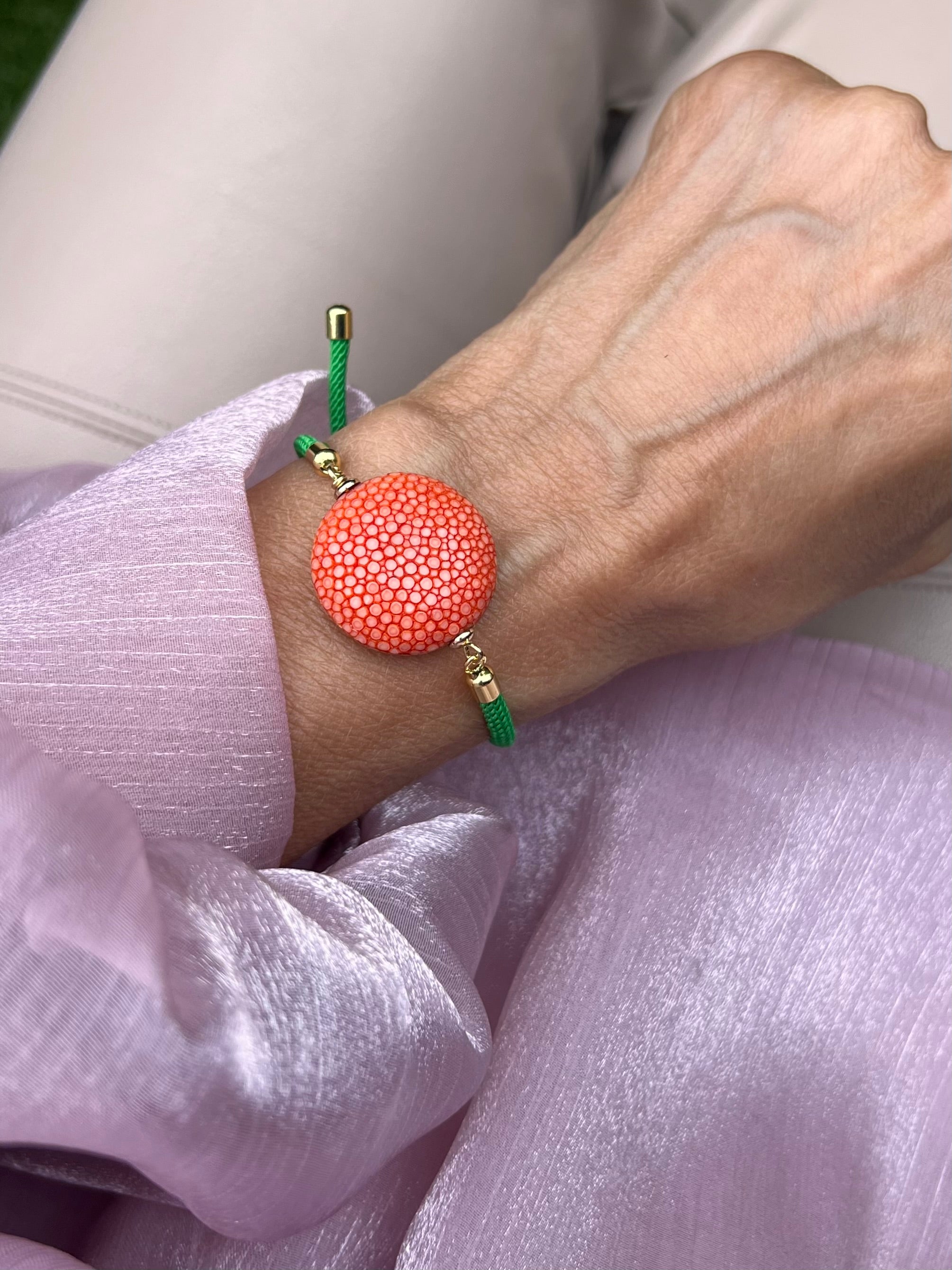 Grünes Armband mit orange Perle und goldene Details. Am Handgelenk fotografiert auf rosa Stoff.