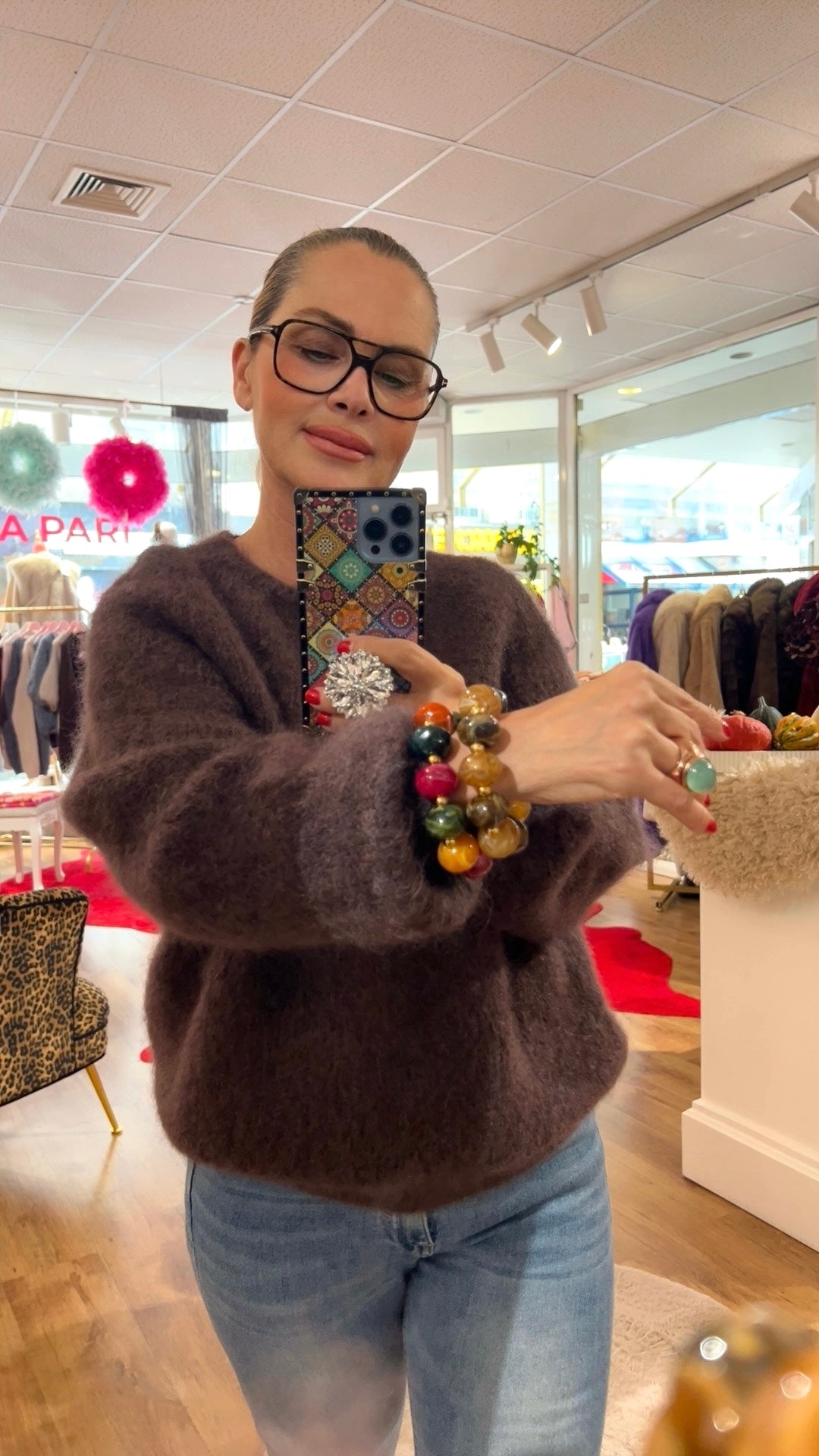 Frau trägt dunkelbraunen Mohairpulli, dazu mehrere bunte Armbänder am Handgelenk und einen großen Ring mit auffälliger Brille und in einer Boutique fotografiert