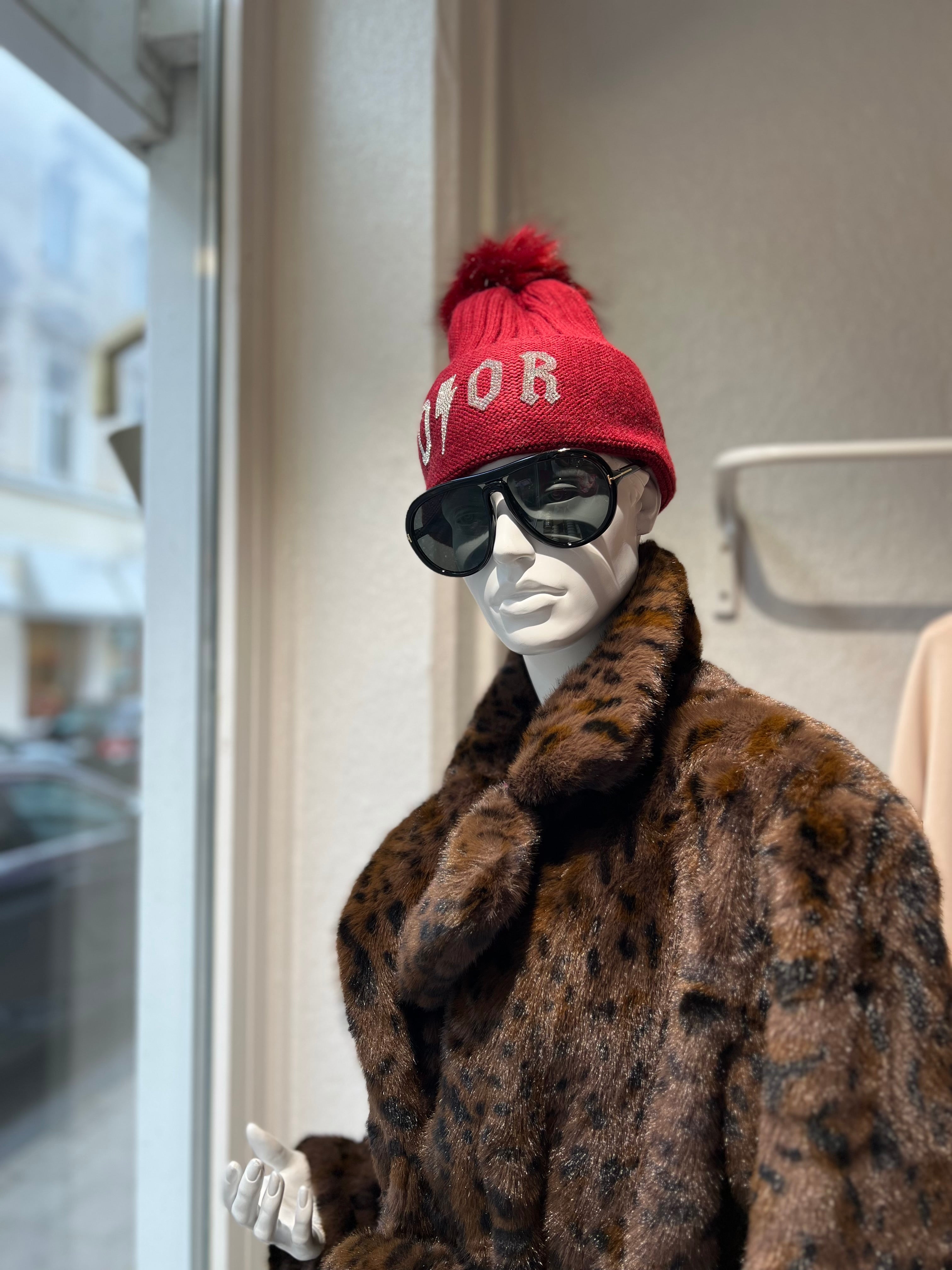 Brauner Kunstpelzmantel mit Leopardenmuster, rote Strickmütze und schwarze Sonnenbrille.