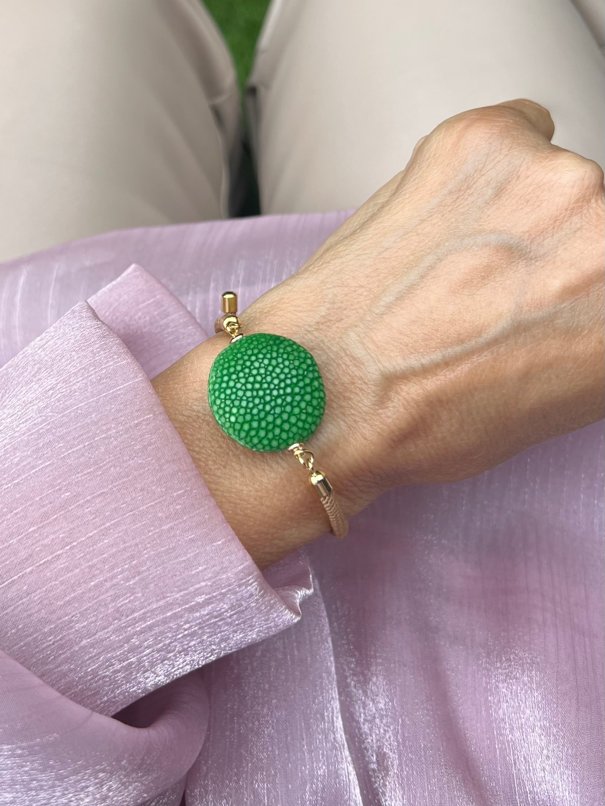 Beiges Armband mit grüner Perle und goldene Details. Am Handgelenk auf rosa Stoff fotografiert.