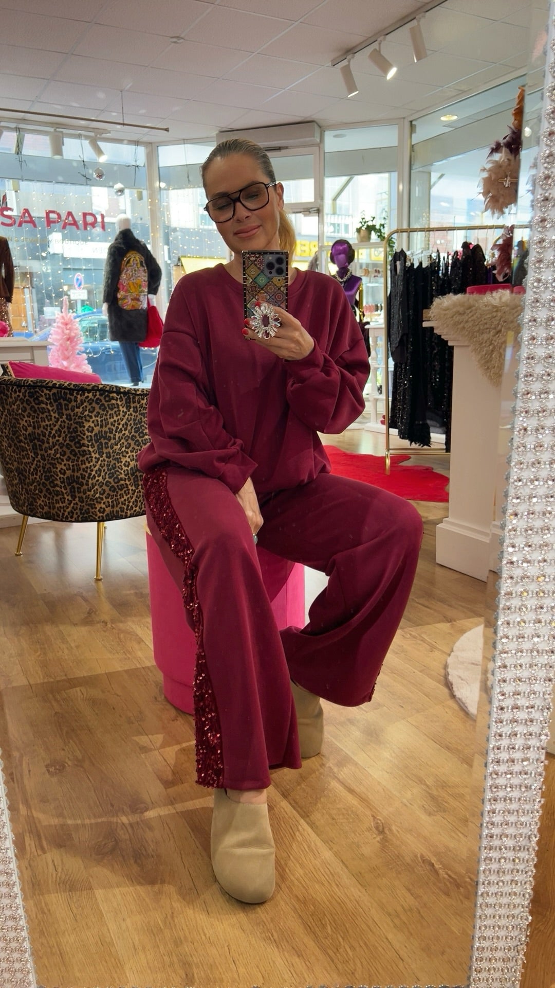 Person trägt ein Burgundy Zweiteiler-Set mit weitem Bein und Paillettenstreifen an der Seite, sitzt auf einem pinken Hocker in einem Boutique-Raum mit Holzfußboden.