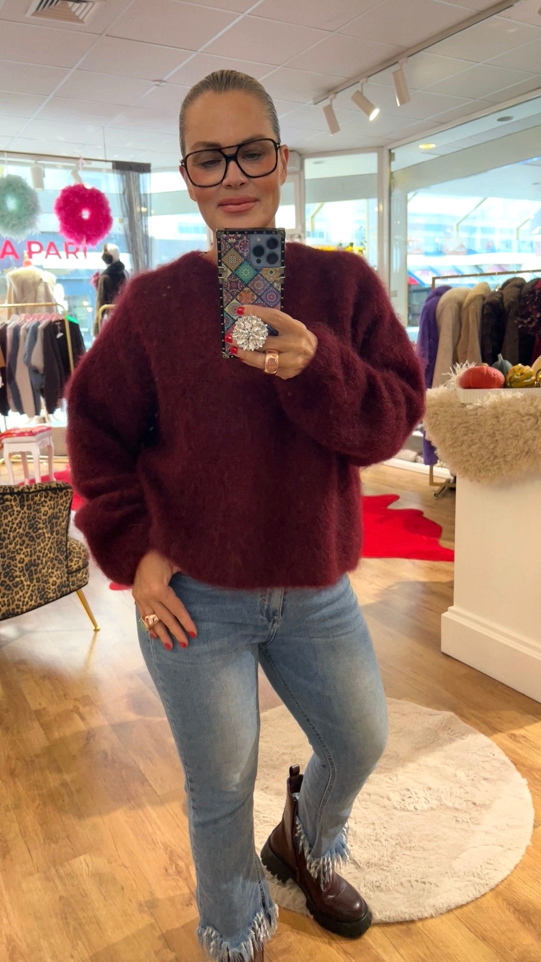 Frau trägt Bordeaux Farben, den Wolle Pulli, dazu eine Jeanshose und braune Stiefel. Sie hält ein Handy in der Hand, fotografiert in einer Boutique.