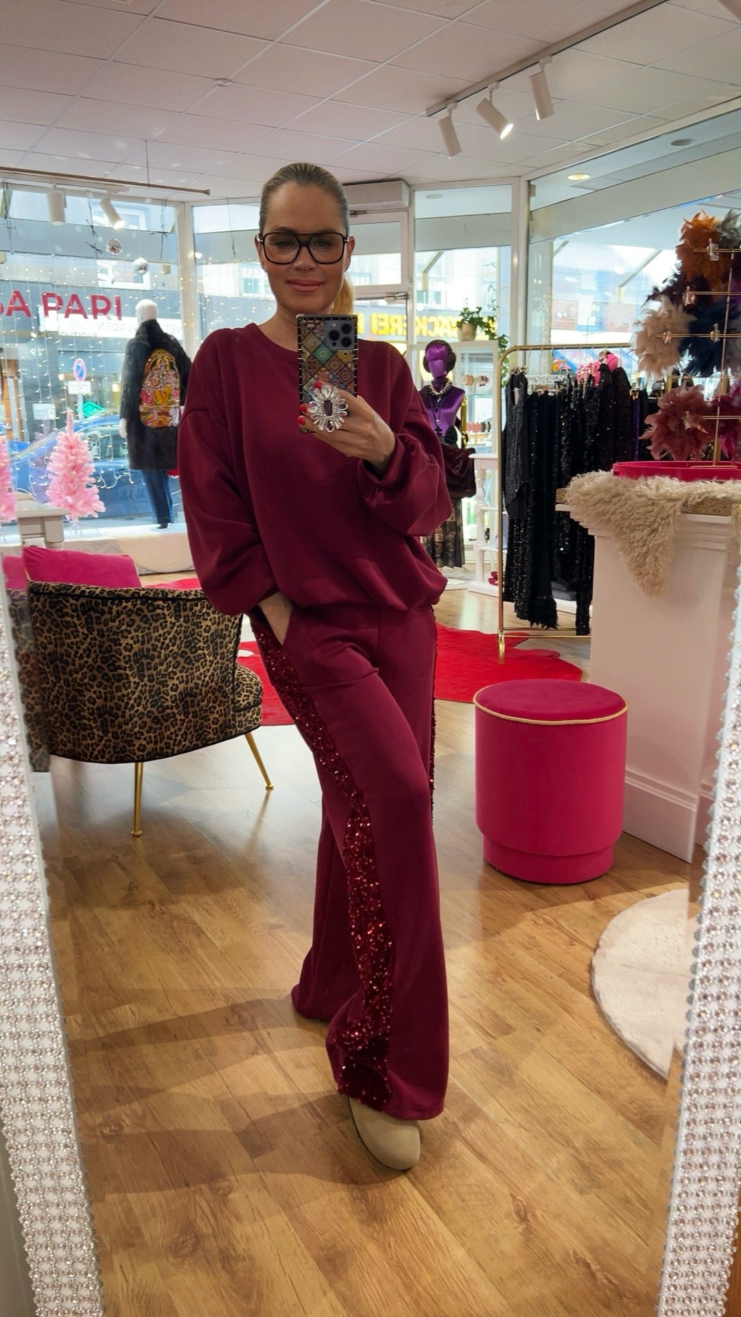 Person steht in einem Burgundy Zweiteiler mit funkelndem Paillettenstreifen an der Seite, in einer Boutique mit Spiegel und Dekoration.
