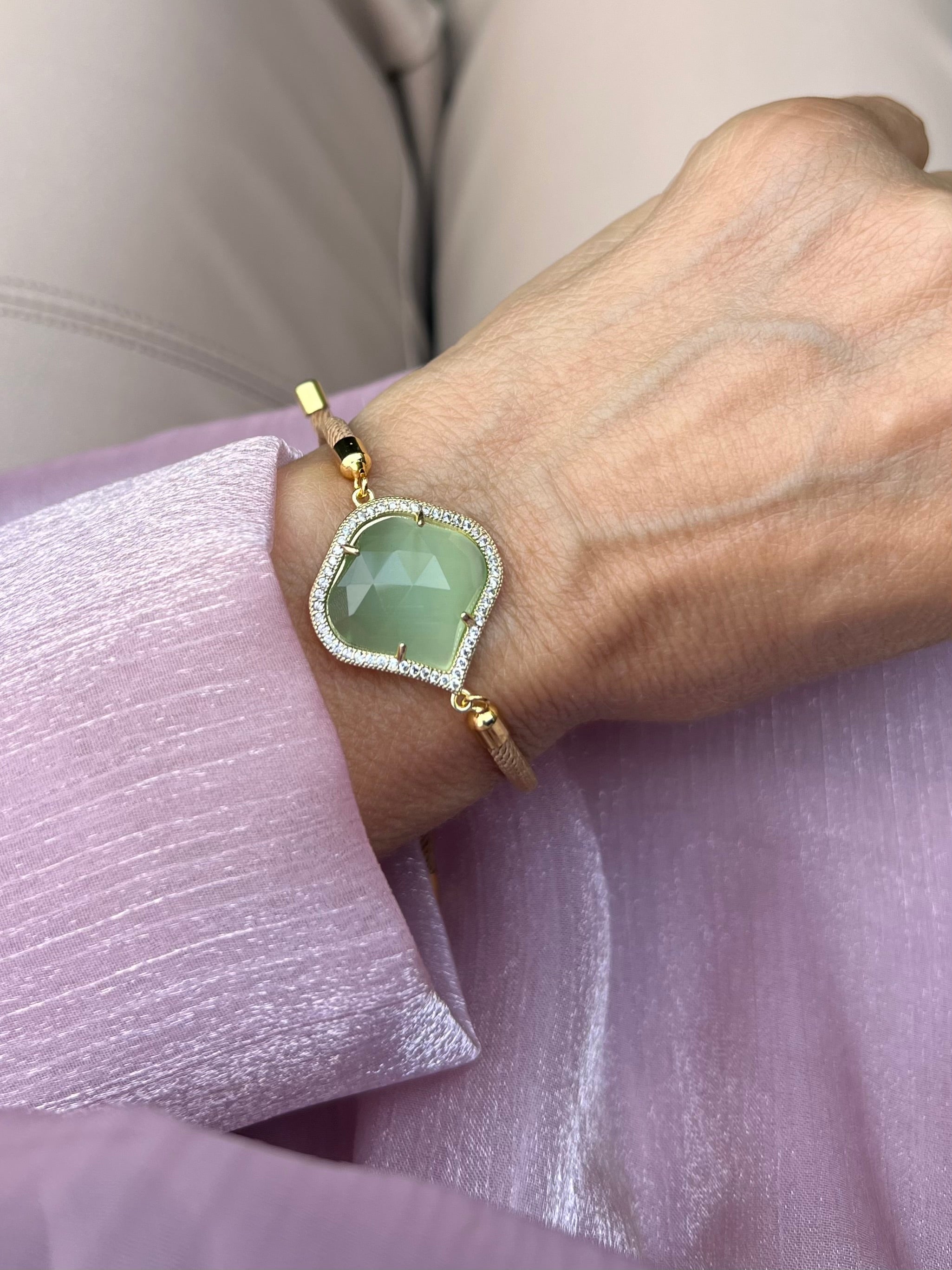 Beiges Armband mit grünem Schmuckstein an Hand getragen. Auf rosa Stoff fotografiert.
