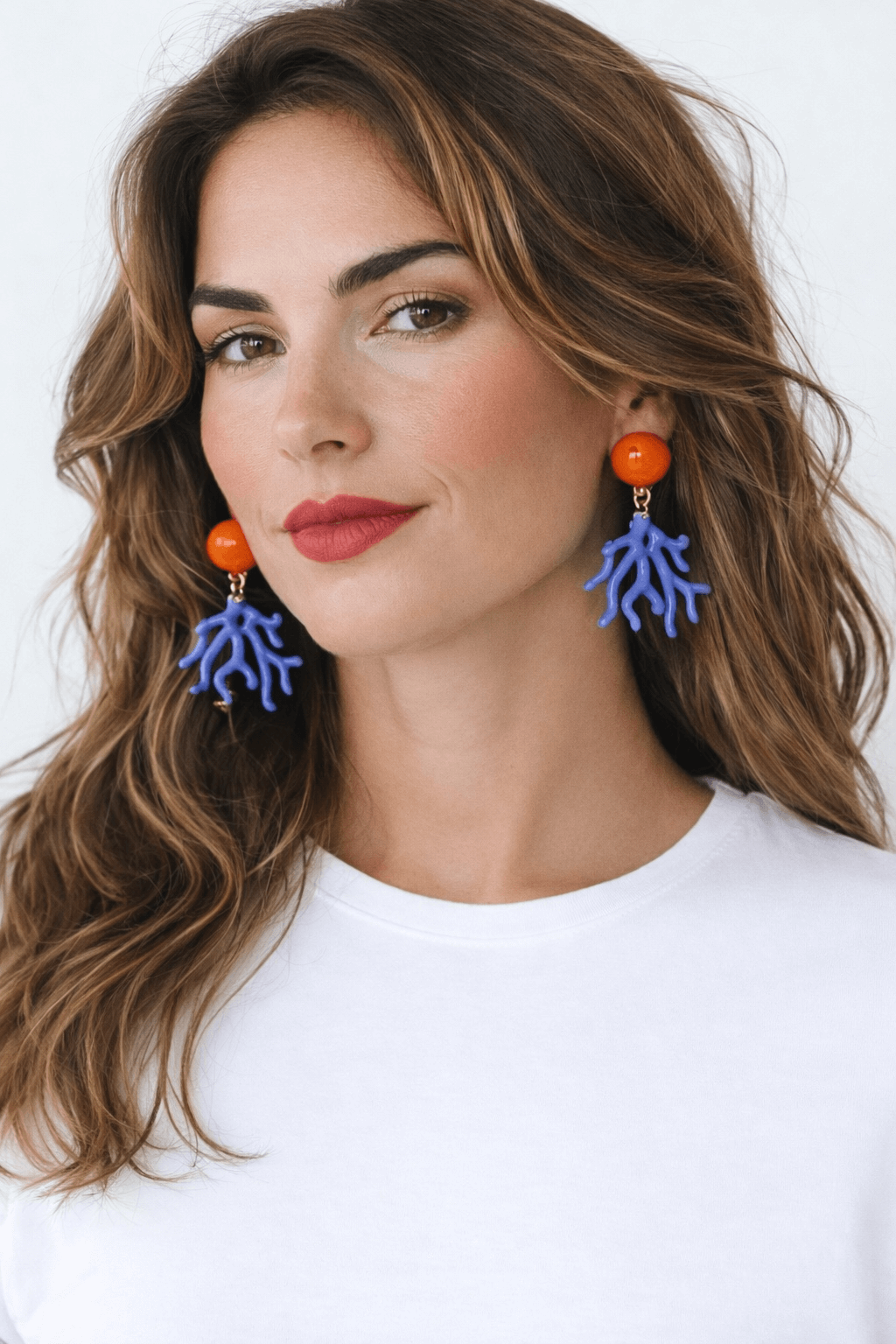Frau mit braunen, welligen Haaren vor hellem Hintergrund, die blaue Clip-Ohrringe in Korallenform mit orangefarbenem, rundem Clipselement trägt.