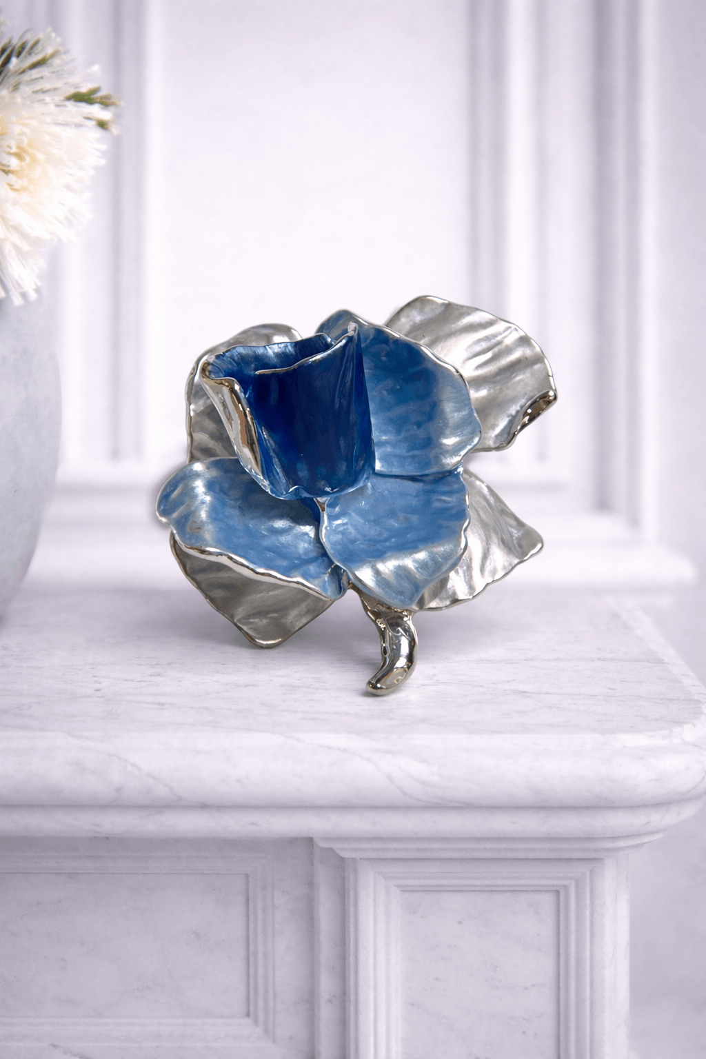 Magnetbrosche in Blütenform mit emaillierten Blütenblättern in Blau sowie silberner Einfassung, fotografiert vor hellem Hintergrund.

