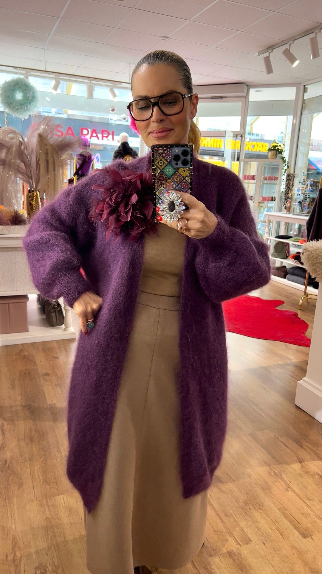 Person steht in einem Boutique-Innenraum und fotografiert sich im Spiegel. Sie trägt einen langen, flauschigen lila Strickcardigan mit weiten Ärmeln über einem beigefarbenen Outfit. An der Schulter ist eine große, dunkelrot-lila Stoff-Blume angebracht. Die Person hält ein Smartphone mit bunter Hülle und funkelndem Ring-Griff. Im Hintergrund sind Regale, Modeartikel, Dekoration und Holzfußboden zu sehen.