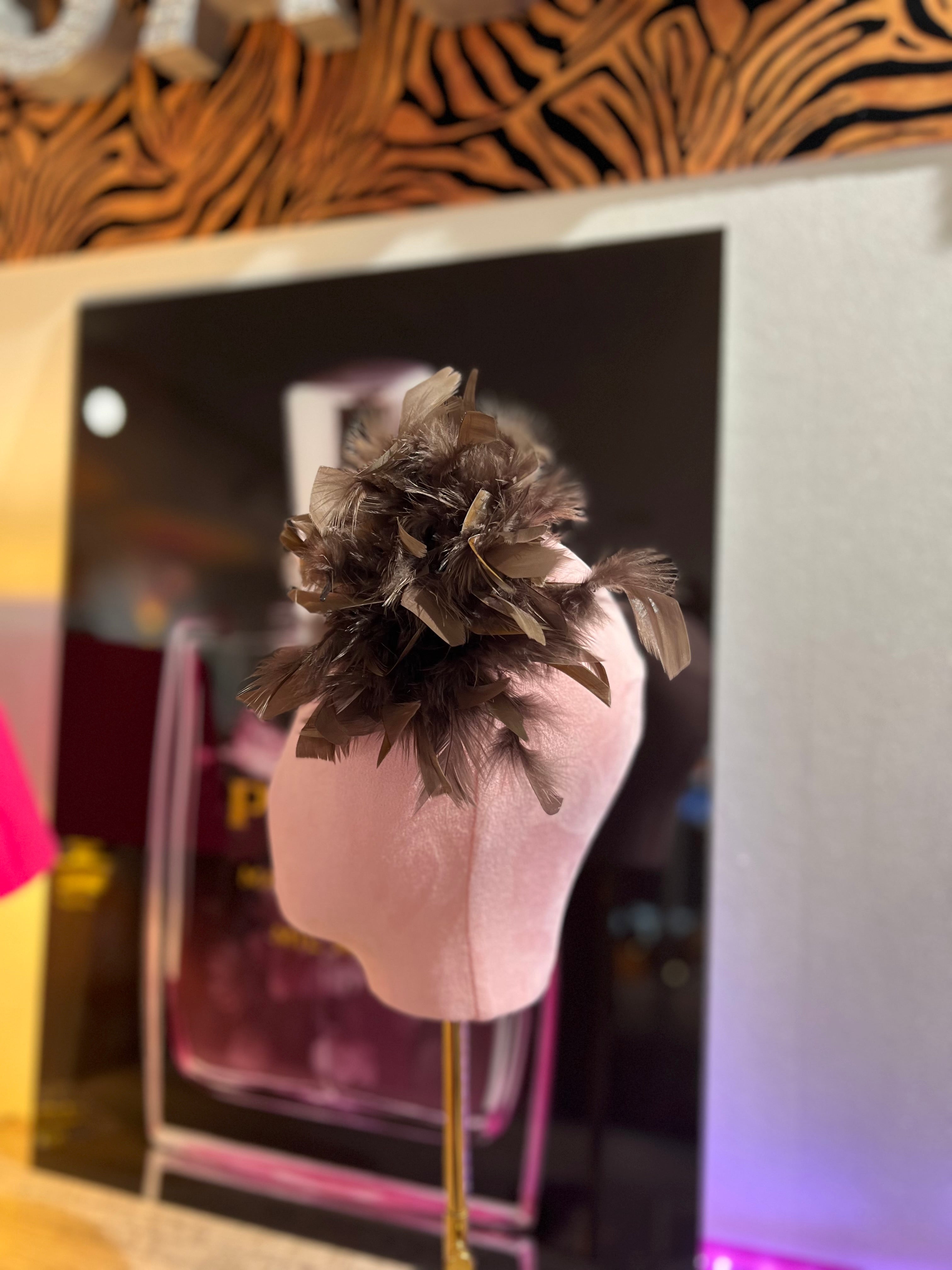 Federbrosche mit großer Blüte aus braunen Federn, befestigt auf einer rosa Stoffpuppe vor dekorativen Hintergrund mit Parfumflakon Motiv.