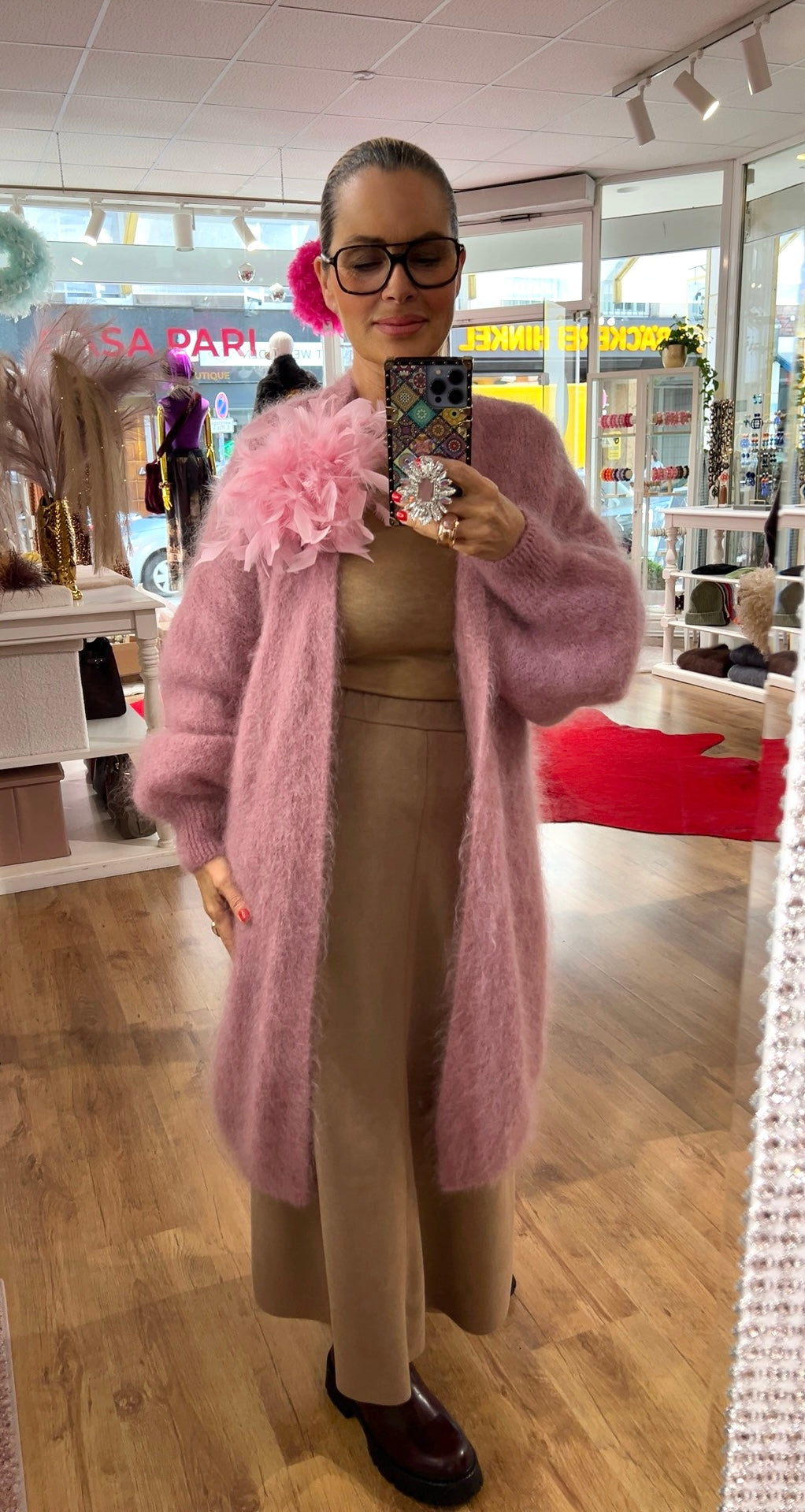 Person steht in einem Boutique-Innenraum und fotografiert sich im Spiegel. Sie trägt einen langen, flauschigen rosafarbenen Strickcardigan mit weiten Ärmeln über einem beigefarbenen Outfit und dunklen Schuhen. Eine große rosa Stoff-Blume an der Schulter befestigt. Die Person hält ein Smartphone mit bunter Hülle und funkelndem Ring-Griff. Im Hintergrund sind Regale, Modeartikel und Holzfußboden zu sehen.