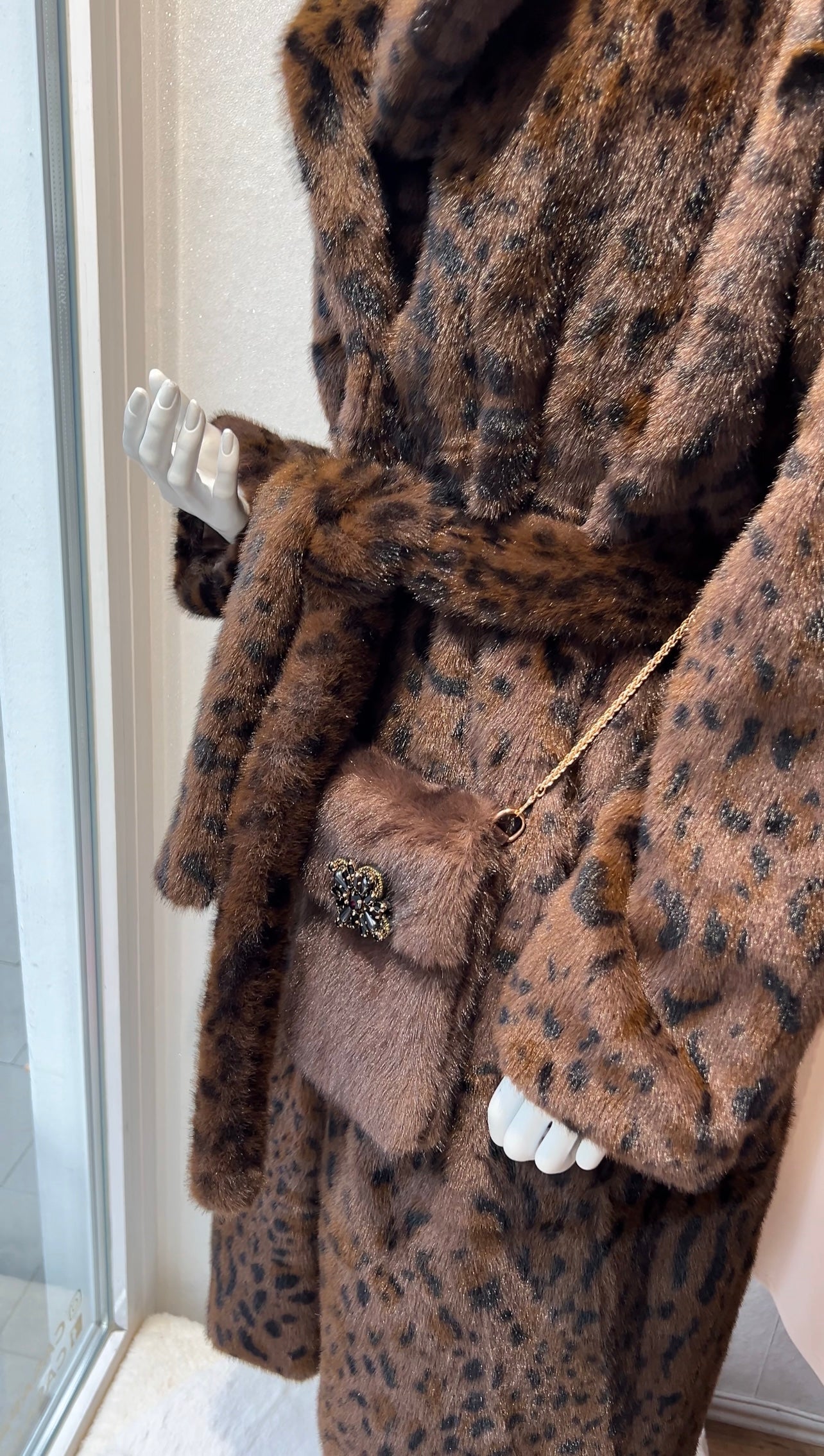 Brauner Kunstpelzmantel mit Leopardenmuster auf einer Schaufensterpuppe, dazu eine kleine Umhängetasche mit goldener Ketten und Brosche.