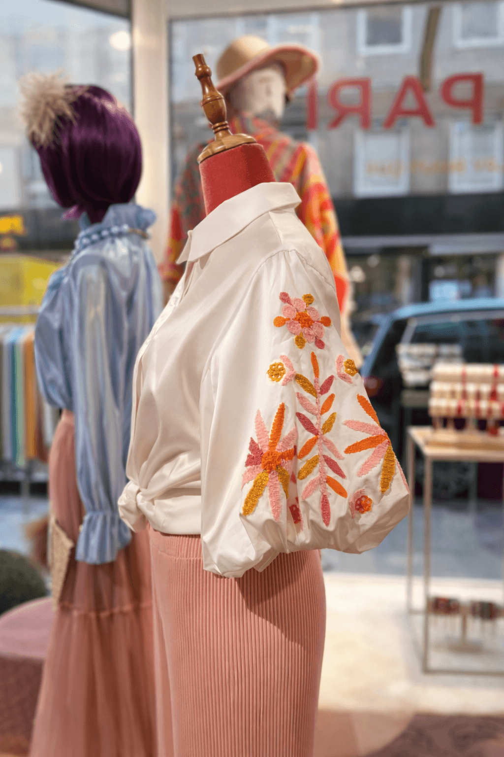 Weiße Damenbluse mit Hemdkragen und Puffärmeln, auf dem rechten Ärmel florale Stickerei in Rosa-, Pink-, Orange- und Goldtönen, kombiniert mit einer rosafarbenen Plisseehose auf einer Schneiderpuppe im Schaufenster einer Boutique.