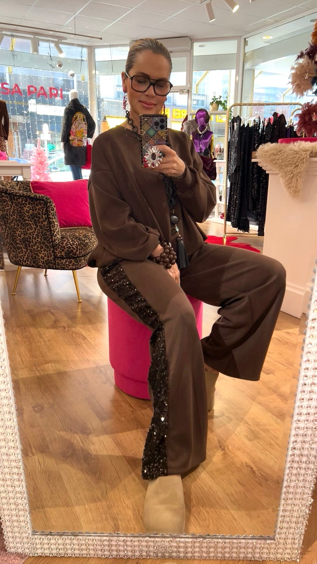 Person trägt ein braunes Zweiteiler-Set mit lockerem Pullover und weitem Bein mit Paillettenstreifen, sitzt entspannt in einer Boutique vor einem Spiegel.