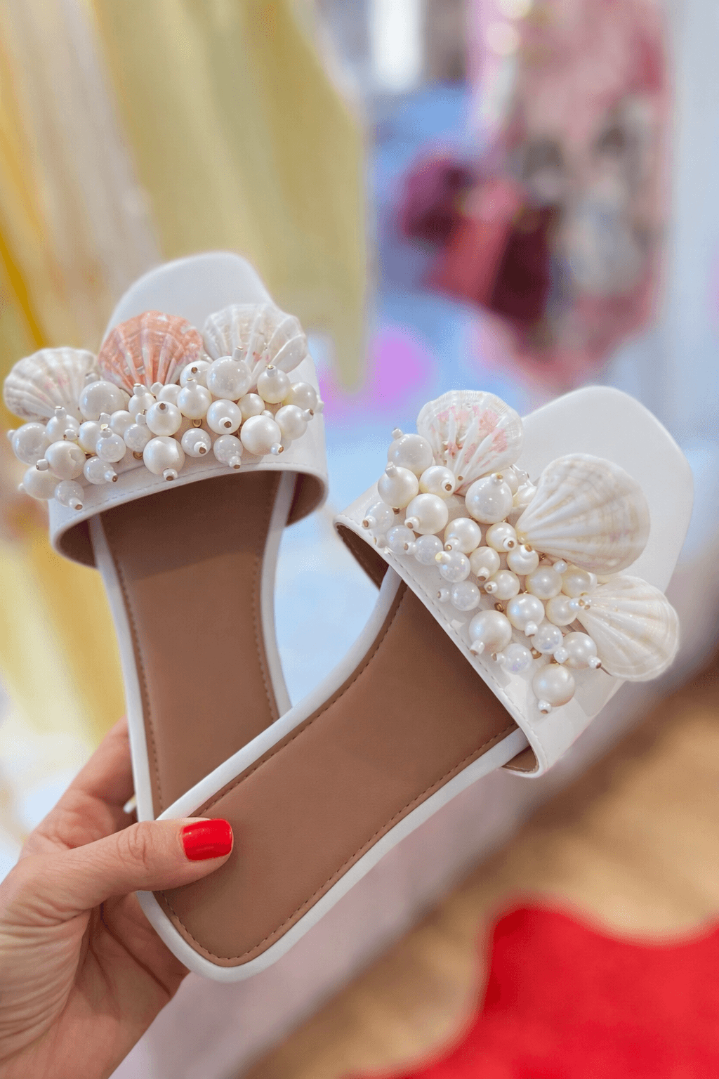 Hand hält ein Paar weiße Sandalen mit Muscheln und Perlen auf dem Riemen; im Hintergrund unscharf ein Boutique-Innenraum mit Kleidung und Teppich.