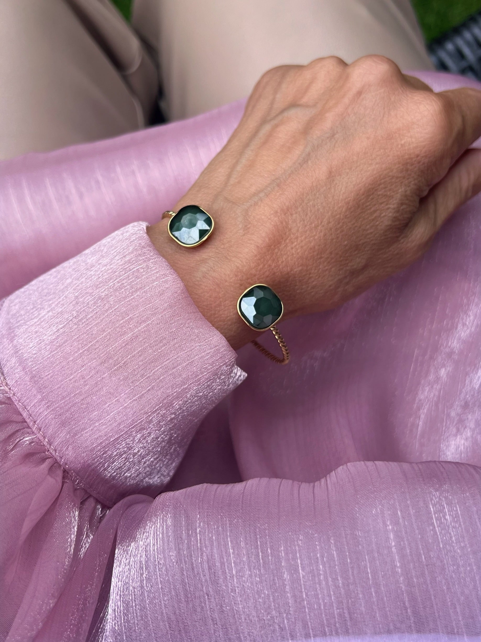 Armreifen in Gold mit grünen Stein, fotografiert an einem Handgelenk auf rosa Stoff.