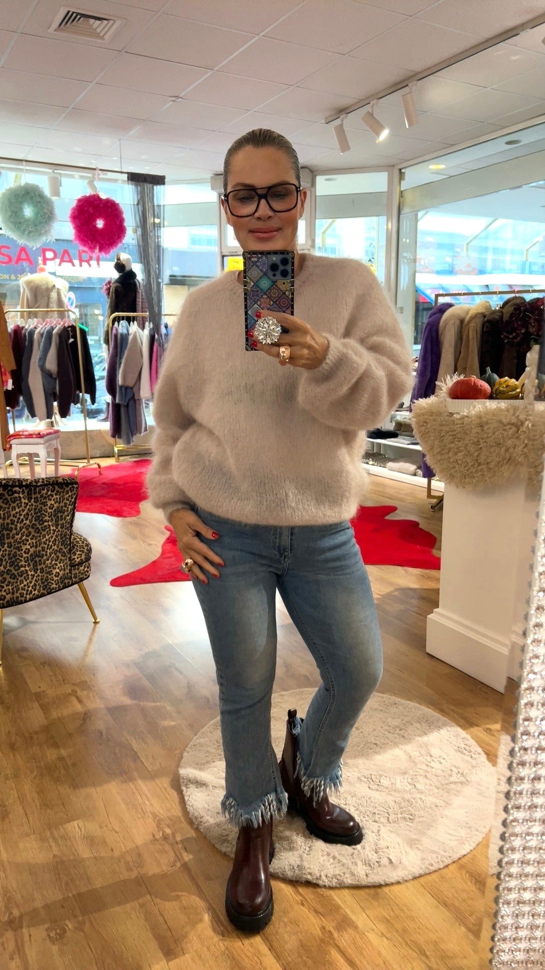 Frau trägt hellen Mohair Pulli, dazu Jeans und braune Boots. Hält ein Handy in der Hand fotografiert in einer Boutique.