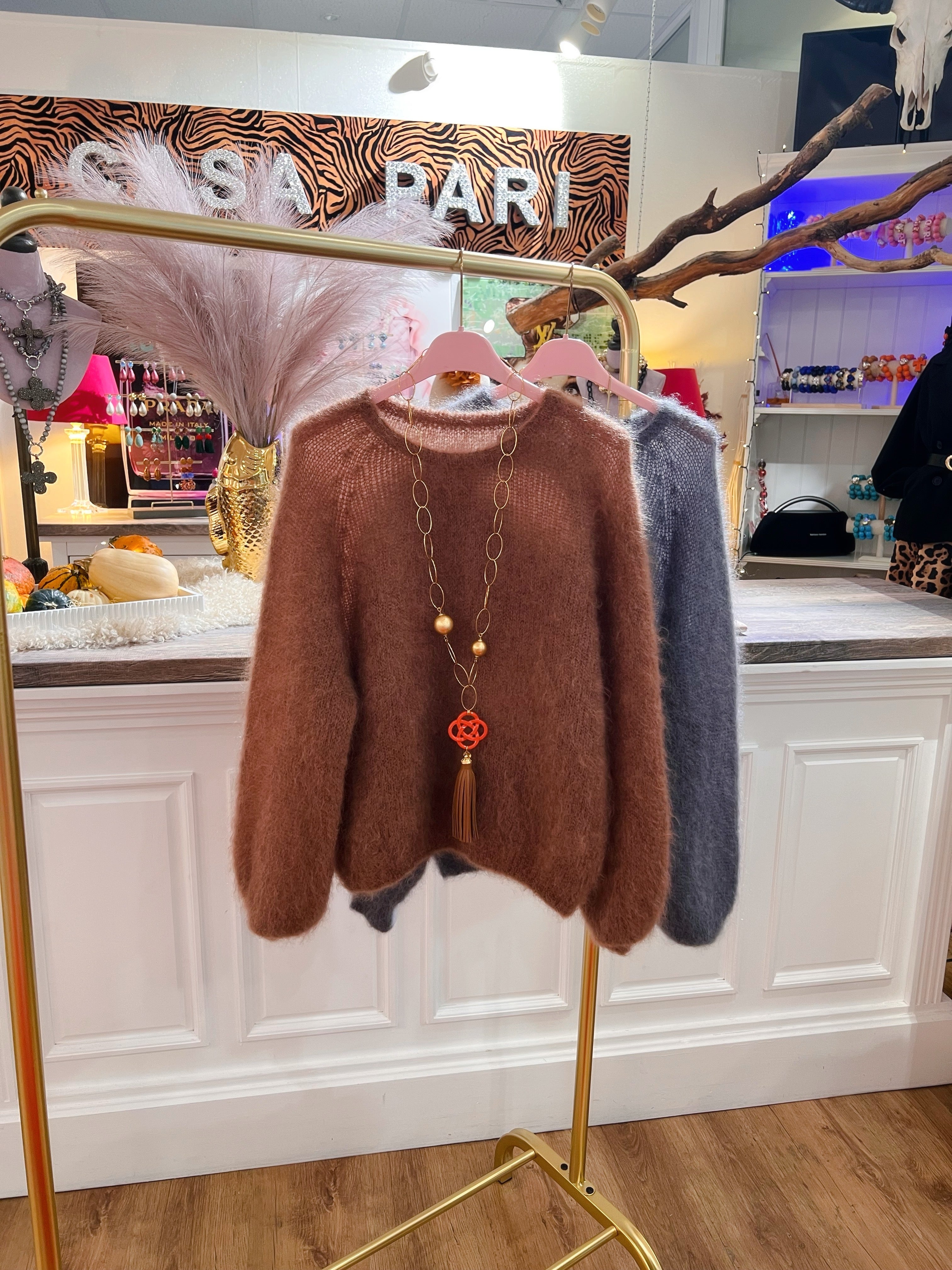 Zwei weiche Pullover aus Kid Mohair in warmen Herbsts Tönen. Karamell mit Kette und Grau. Hängen auf roséfarbenen Kleiderbügel an einer goldenen Stange in einer Boutique.