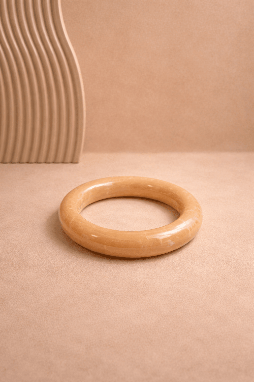 Braun-beiger, glänzender Resin-Armreif liegt mittig auf warmem, sandfarbenem Hintergrund; links im Bild ein geriffeltes Deko-Element, weiches Licht und klare Darstellung.