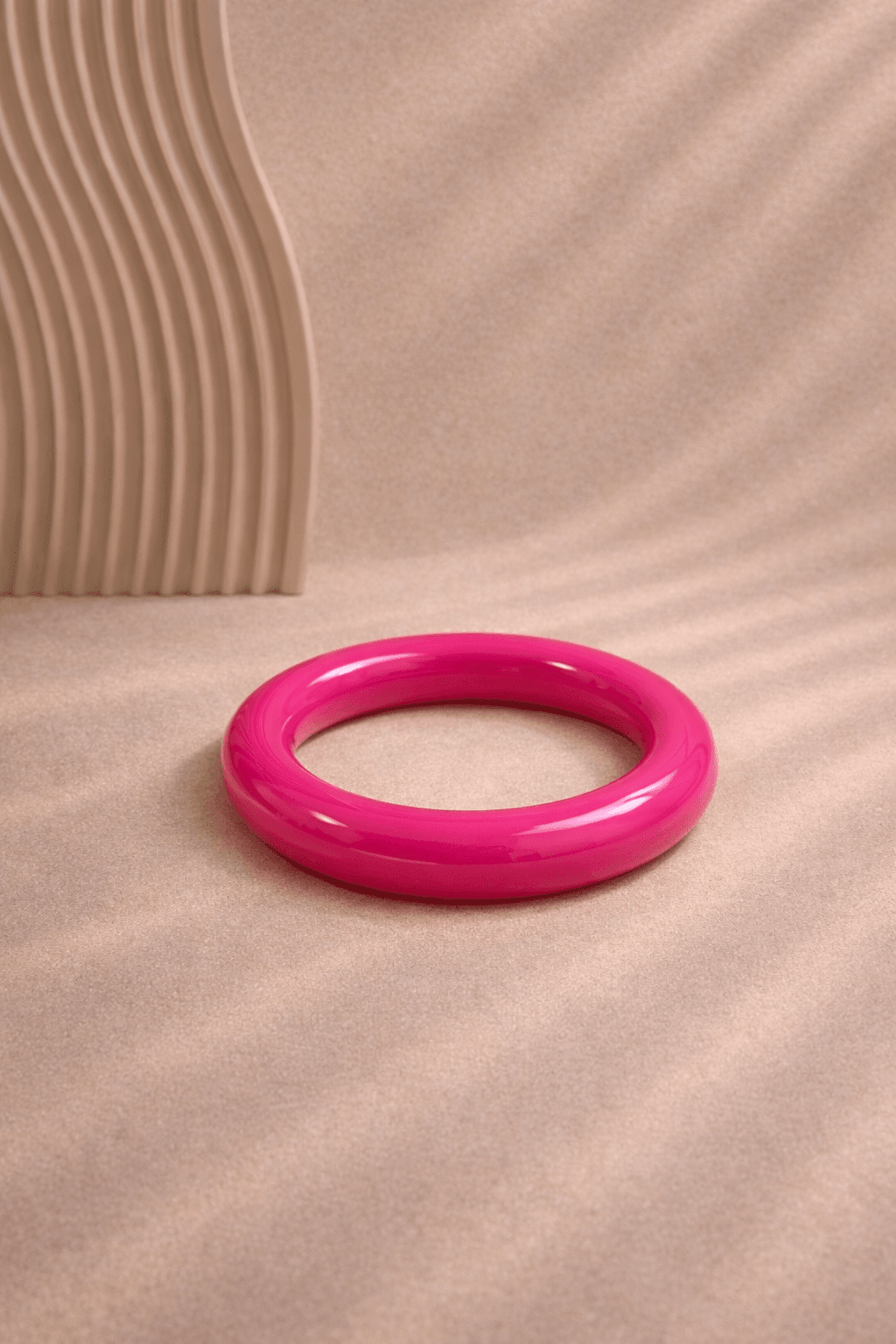 Pinker glänzender Resin-Armreif liegt mittig auf warmem, sandfarbenem Hintergrund; links im Bild ein geriffeltes Deko-Element, weiches Licht und klare Darstellung.