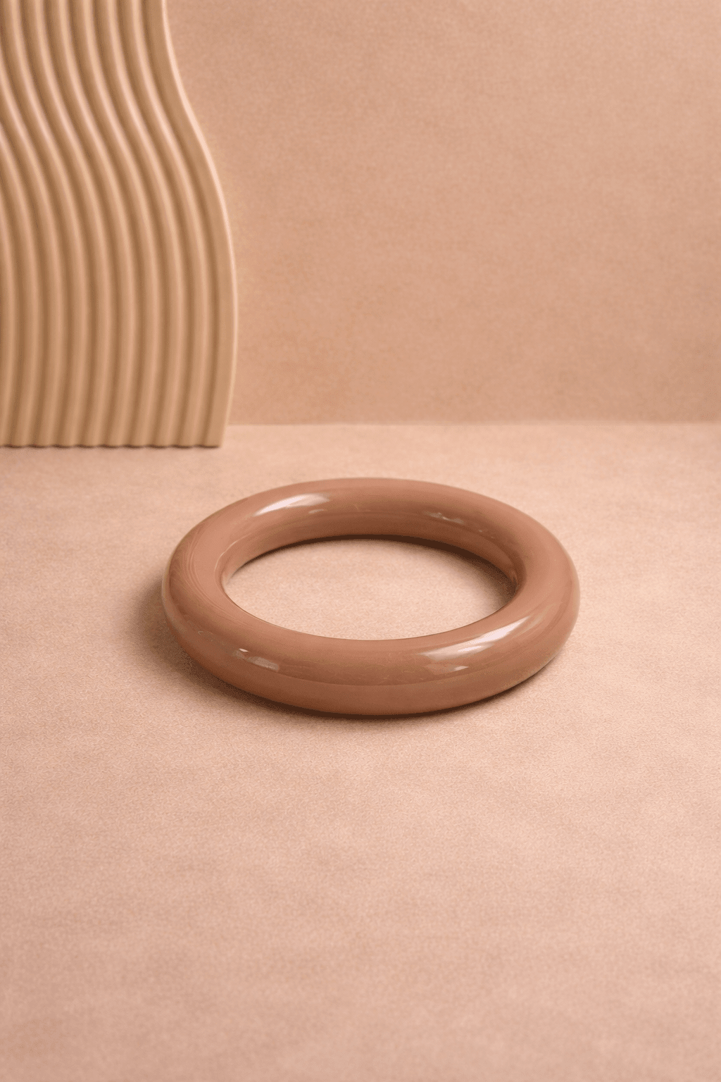 Braun-beiger, glänzender Resin-Armreif liegt mittig auf warmem, sandfarbenem Hintergrund; links im Bild ein geriffeltes Deko-Element, weiches Licht und klare Darstellung.