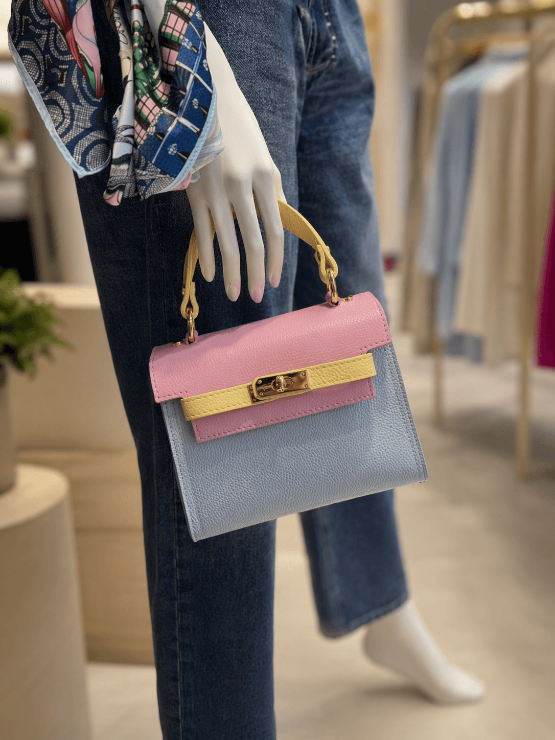 Pastellfarbene Ledertasche mit Henkel und goldfarbenem Verschluss, getragen an der Hand einer Schaufensterpuppe in einer Boutique.