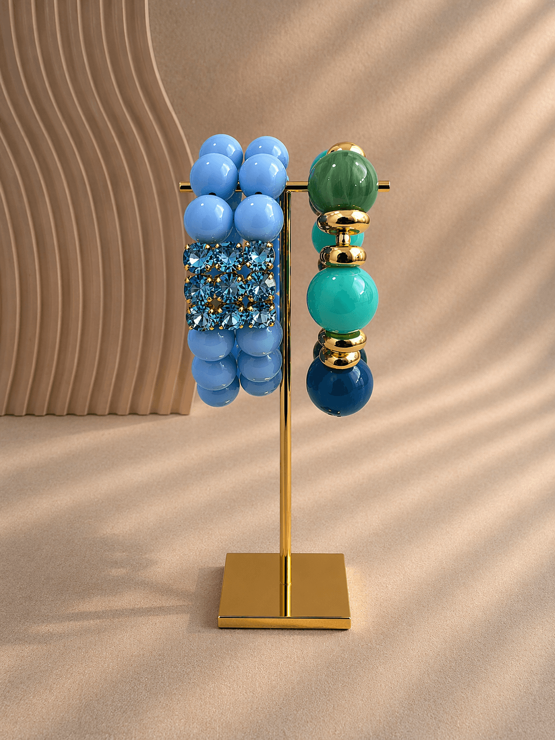 Zwei farbige Armbänder aus großen Resinaperlen hängen an einem goldenen Ständer; links doppelreihig in Hellblau mit Strassdetail, rechts einreihig in Grün-, Türkis- und Blautönen mit goldenen Perlen.