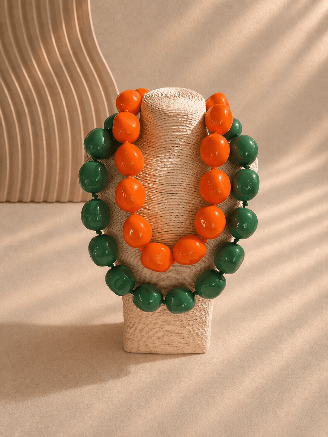 Zwei große Perlenketten in Orange und Grün hängen auf einem beigen, geflochtenen Schmuckständer vor hellem Hintergrund.