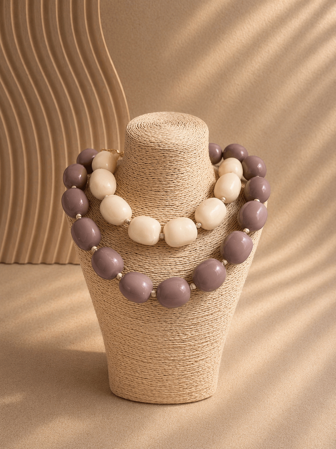 Zwei große Perlenketten in Taupe und Creme hängen auf einem beigen, geflochtenen Schmuckständer vor hellem Hintergrund.