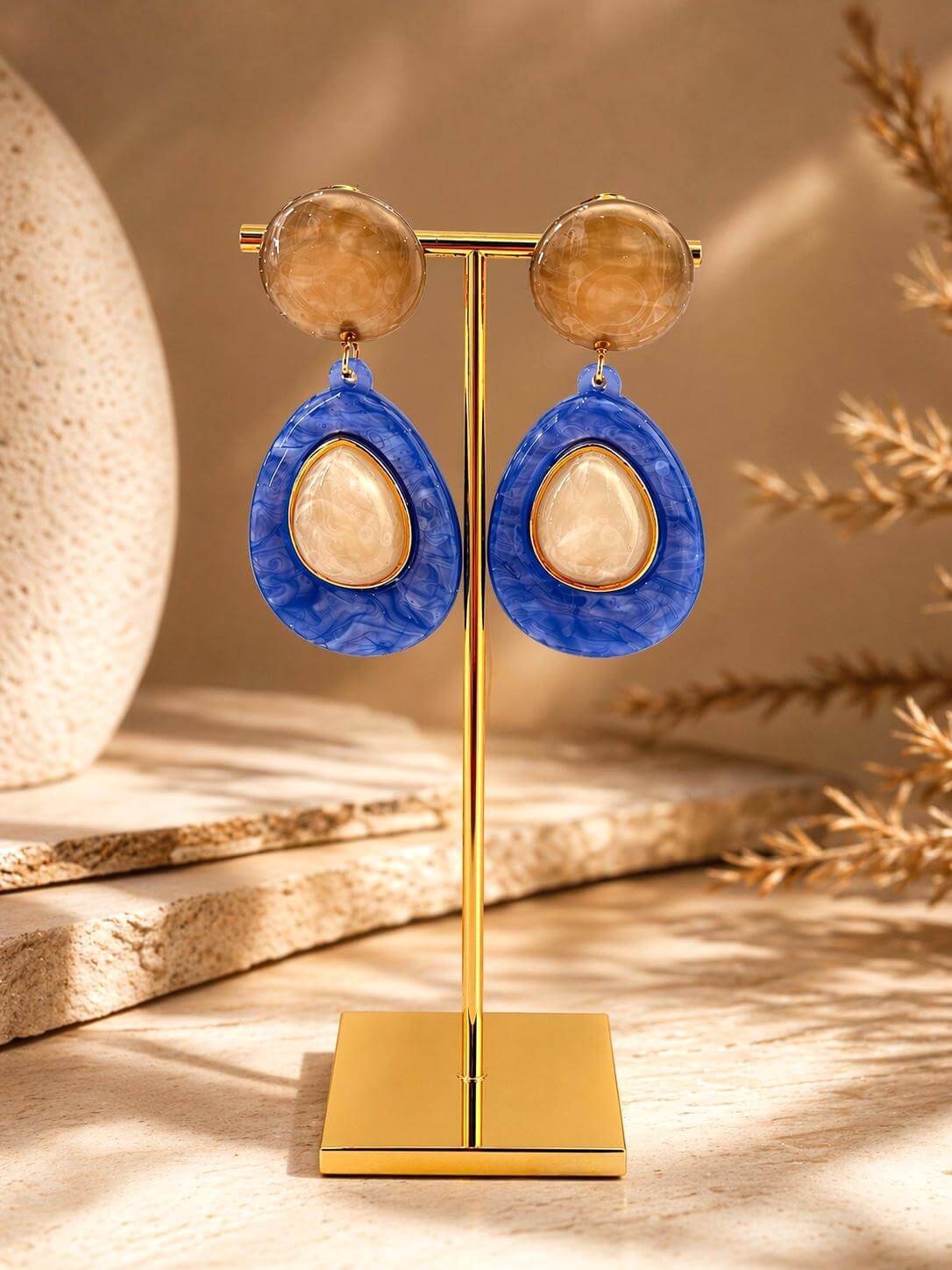 Blau-cremefarbene Statement-Ohrringe aus Resina mit goldfarbenem Rahmen, Tropfenform, zweiteilig, auf goldenem Schmuckständer vor warmem, neutralem 
