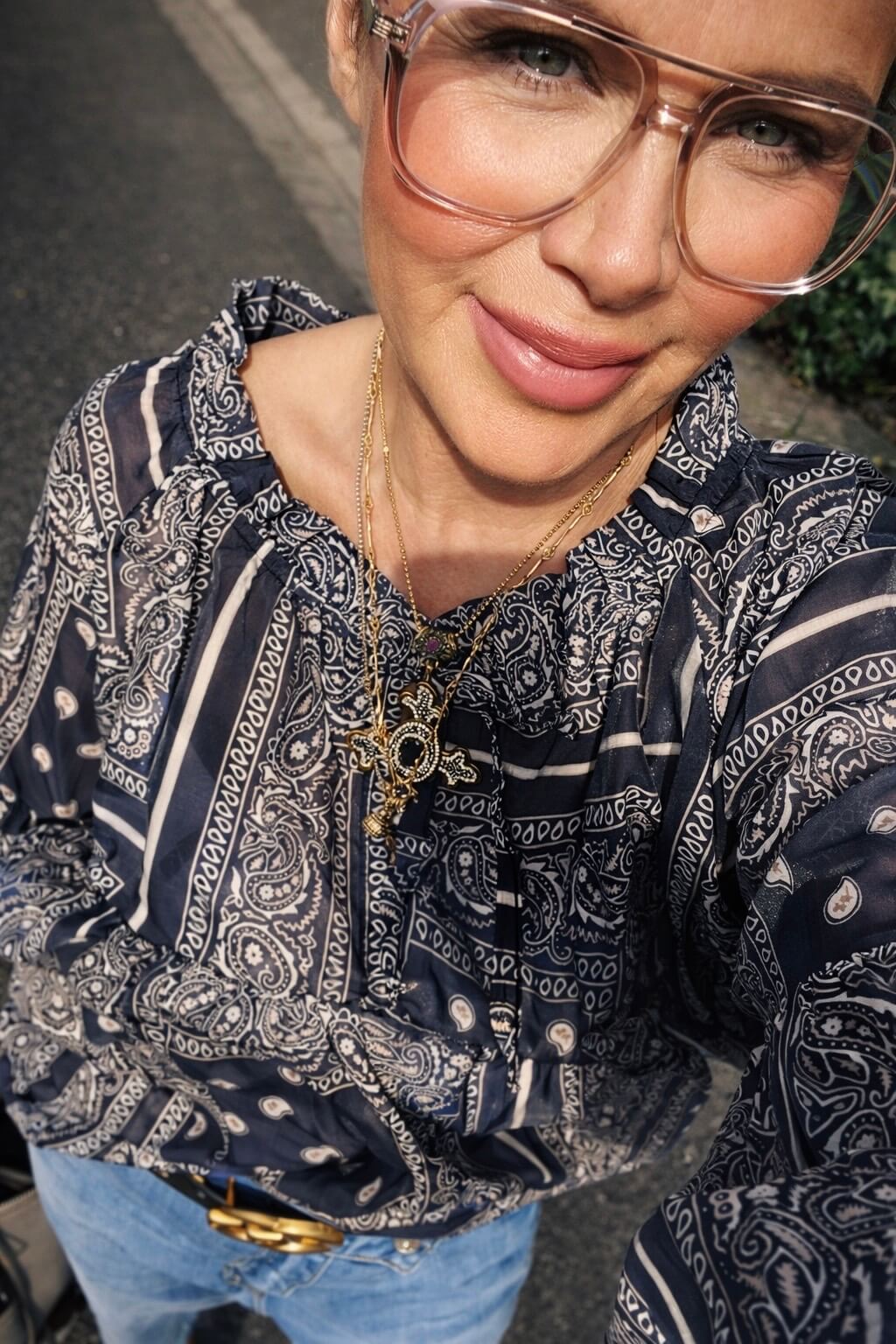 Person im Freien trägt eine dunkelblaue Paisley-Bluse, Jeans und Sandalen sowie eine Brille und mehrere Halsketten, fotografiert aus leicht erhöhter Perspektive bei Sonnenlicht.