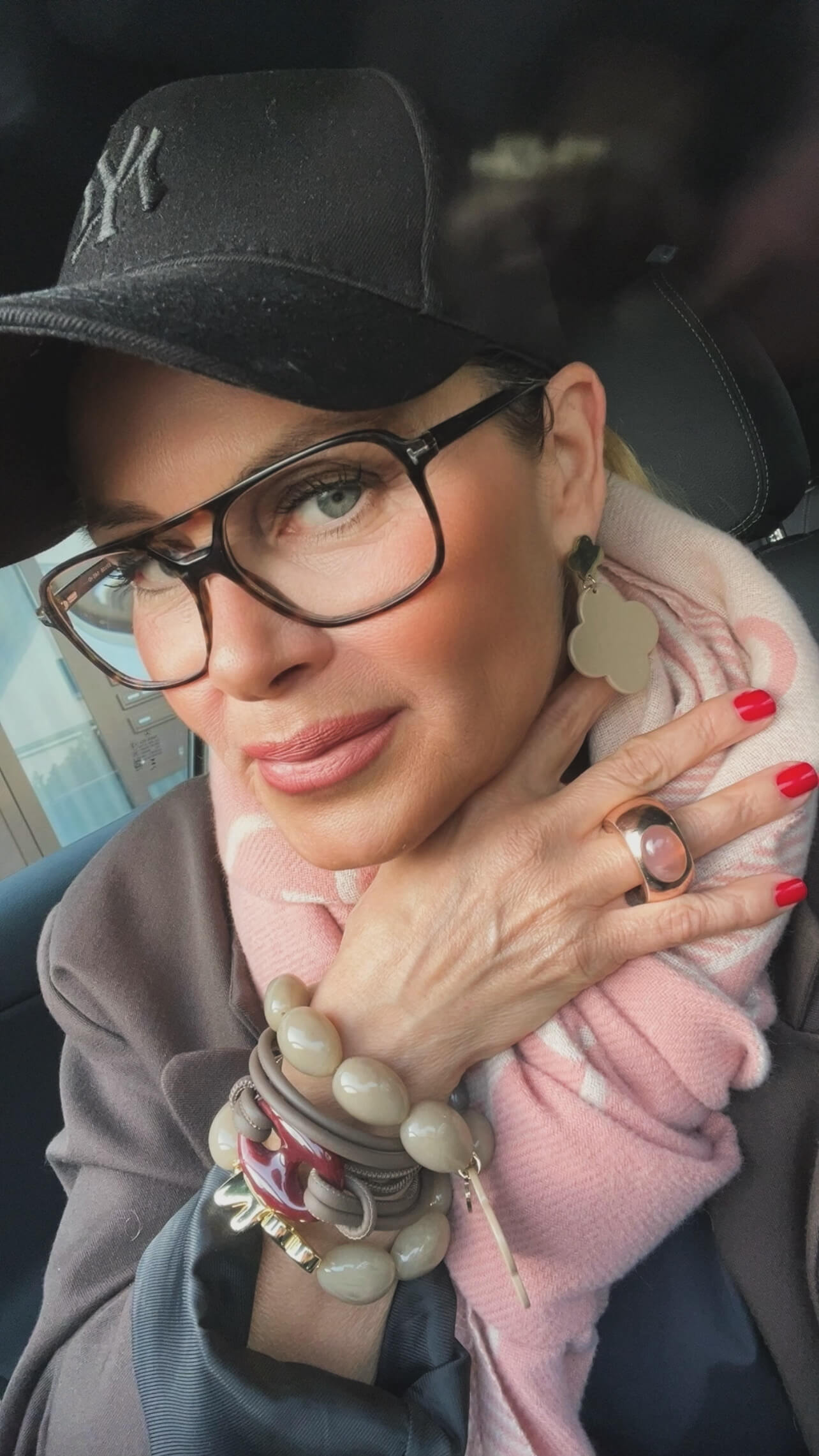 Person mit schwarzer Baseballkappe, Brille und beigefarbenem Kleeblatt-Ohrring. Die Hand trägt mehrere Armbänder und einen Ring mit rundem Stein.