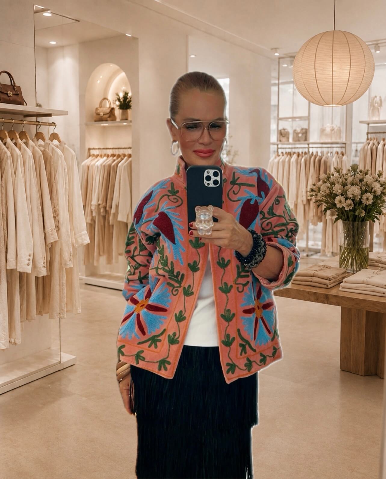 Frau fotografiert sich in einem hellen Boutique-Spiegel. Sie trägt eine rosafarbene, bunt bestickte Jacke mit floralem Muster über einem weißen Oberteil und einem schwarzen Fransenrock. Im Hintergrund sind helle Kleiderstangen, ein Holztisch mit gefalteter Kleidung und ein Blumenstrauß zu sehen.