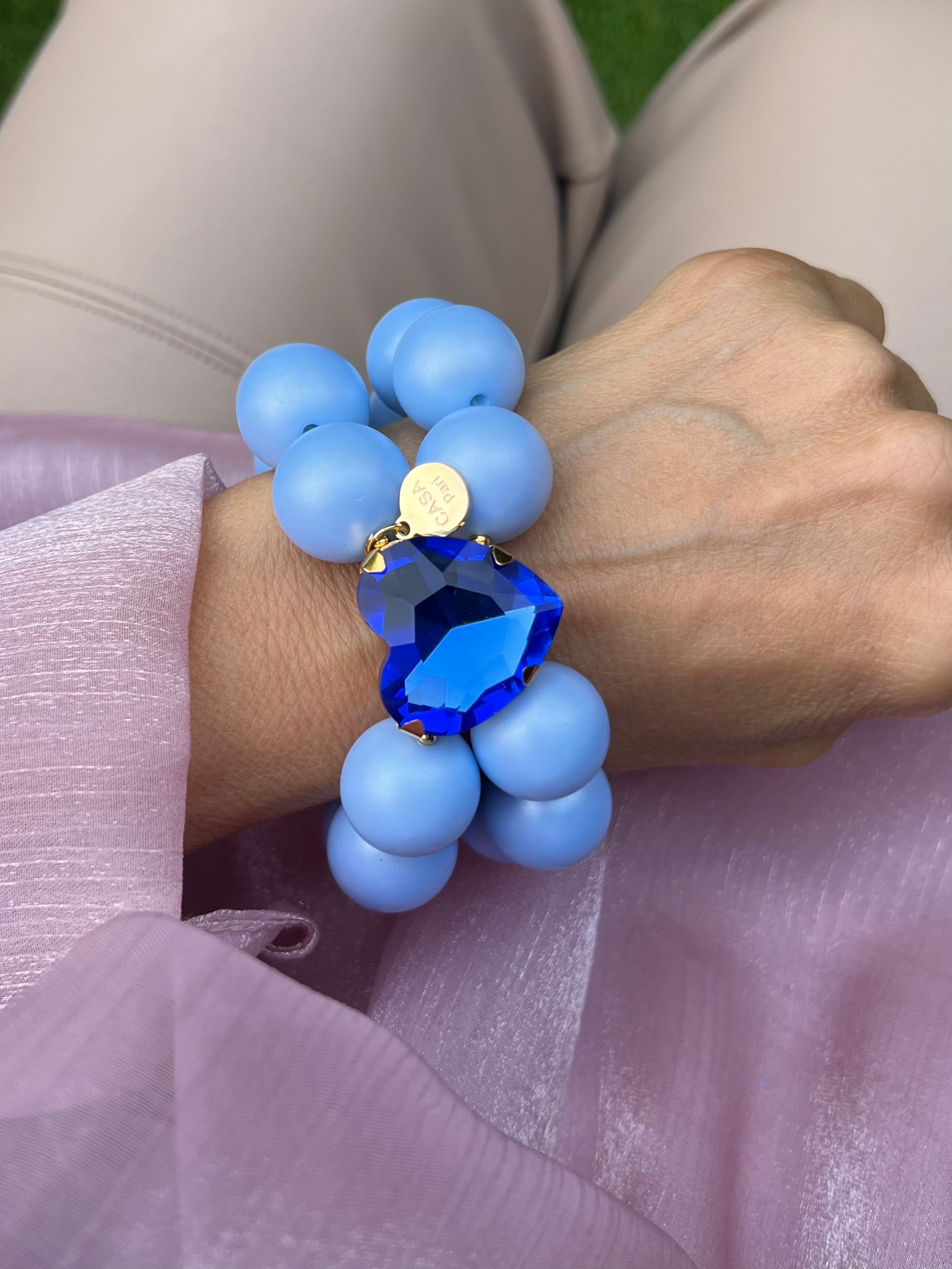 Handgelenk mit blauem Armband aus großen, glänzenden Perlen und einem herzförmigen, tiefblauen Kristall in der Mitte.