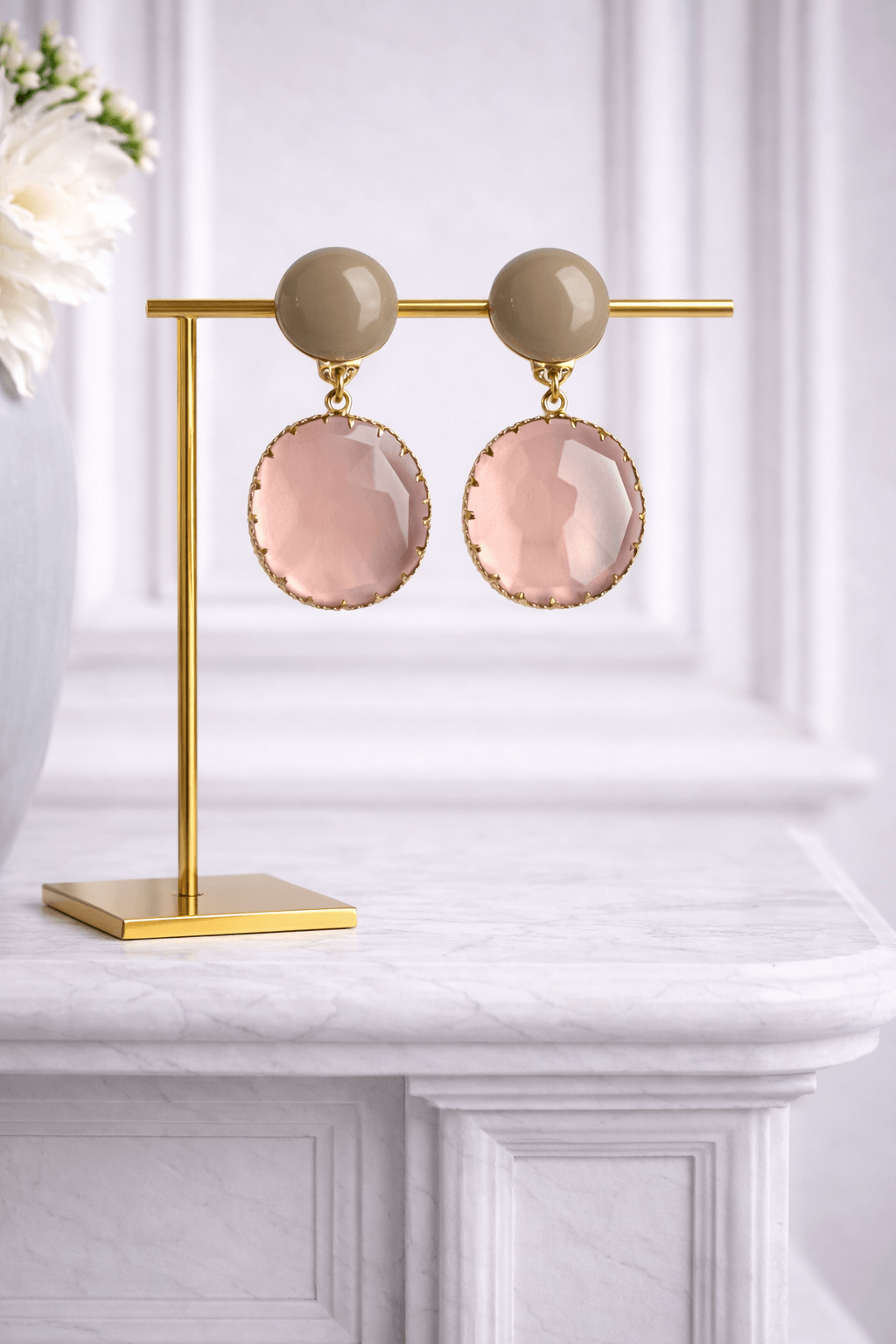 Paar roséfarbene, runde Glas-Ohrringe mit facettiertem Stein und nude-farbenem Stecker, präsentiert auf goldenem Schmuckständer vor hellem Hintergrund.