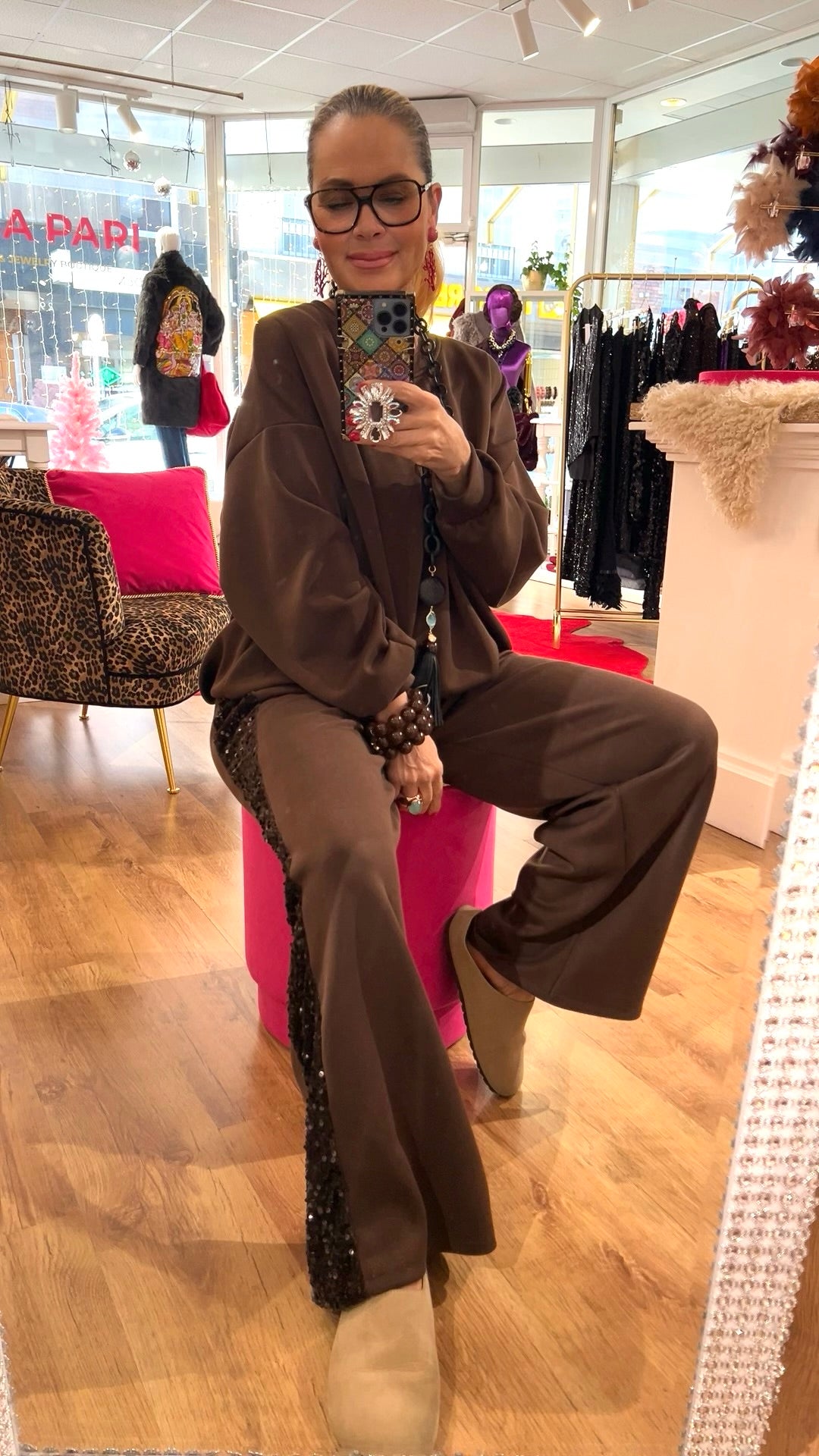 Person trägt ein braunes Zweiteiler-Set mit weitem Bein und Paillettenstreifen an der Seite, sitzt auf einem pinken Hocker in einem Boutique-Raum mit Holzfußboden.