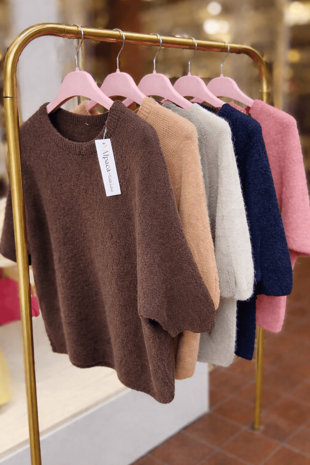 Mehrere kurzärmelige Strickpullover aus Alpaka-Mischung hängen auf rosa Kleiderbügeln an einer goldfarbenen Kleiderstange. Die Pullover sind in verschiedenen Farben angeordnet, darunter Braun, Beige, Creme, Dunkelblau und Rosa. Im Hintergrund ist unscharf das Schaufenster eines Geschäfts zu sehen.