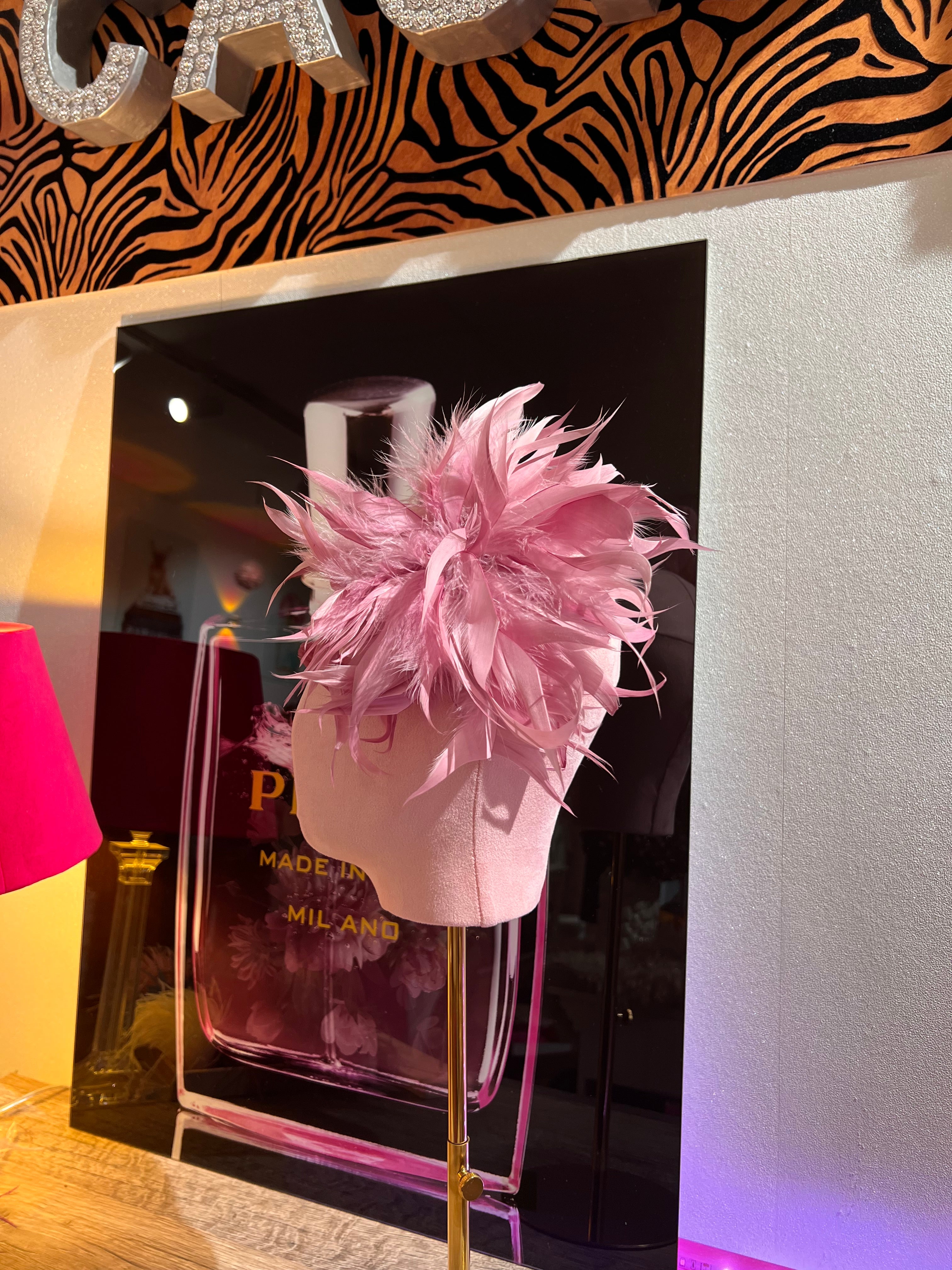 Federbrosche mit großer Blüte aus rosa Federn, befestigt auf einer rosa Stoffpuppe vor dekorativen Hintergrund mit Parfumflakon Motiv.