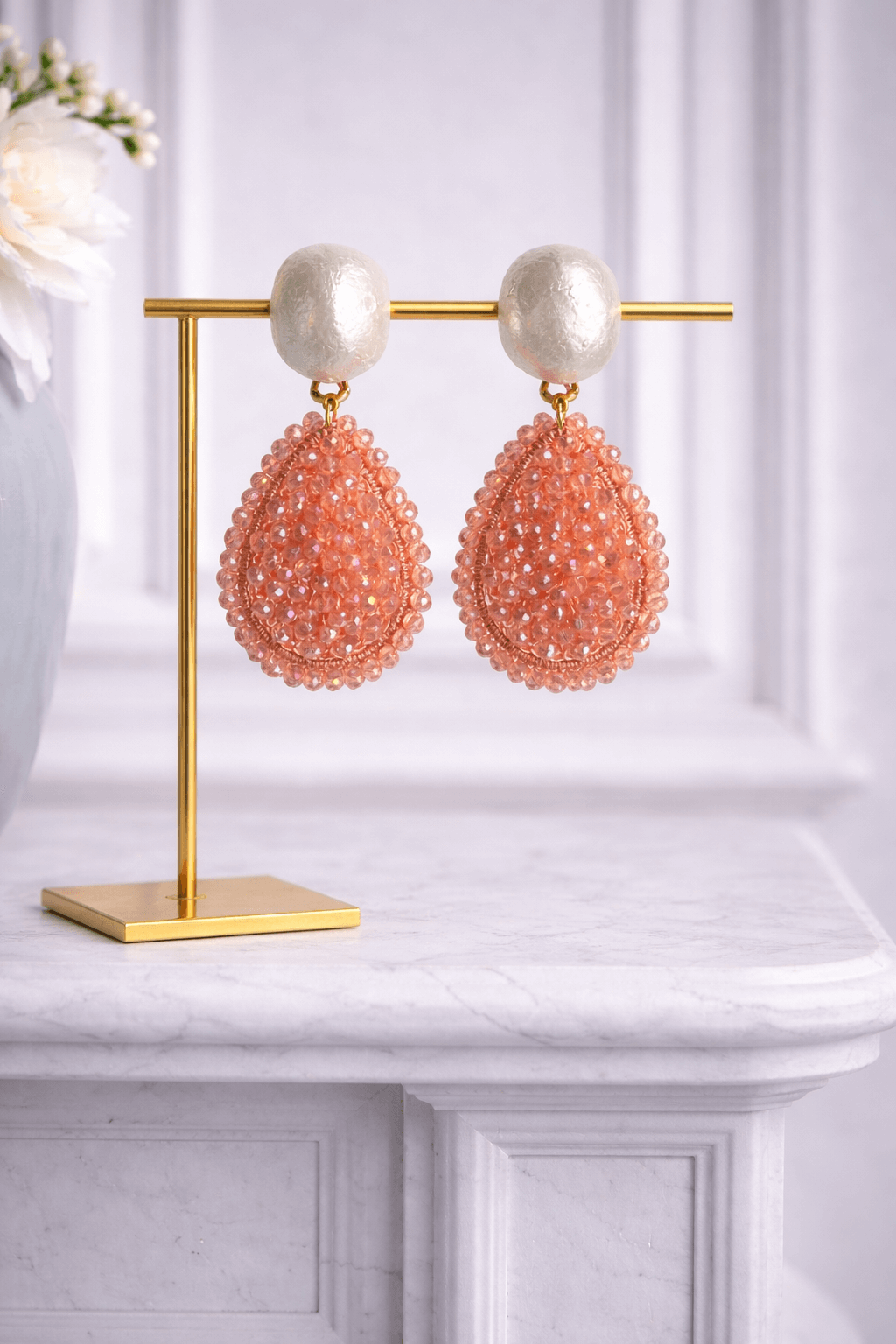 Apricotfarbene Tropfen-Ohrringe mit schimmernden Glasperlen und perlmuttfarbenem Clip auf goldfarbenem Schmuckständer vor hellem Hintergrund.