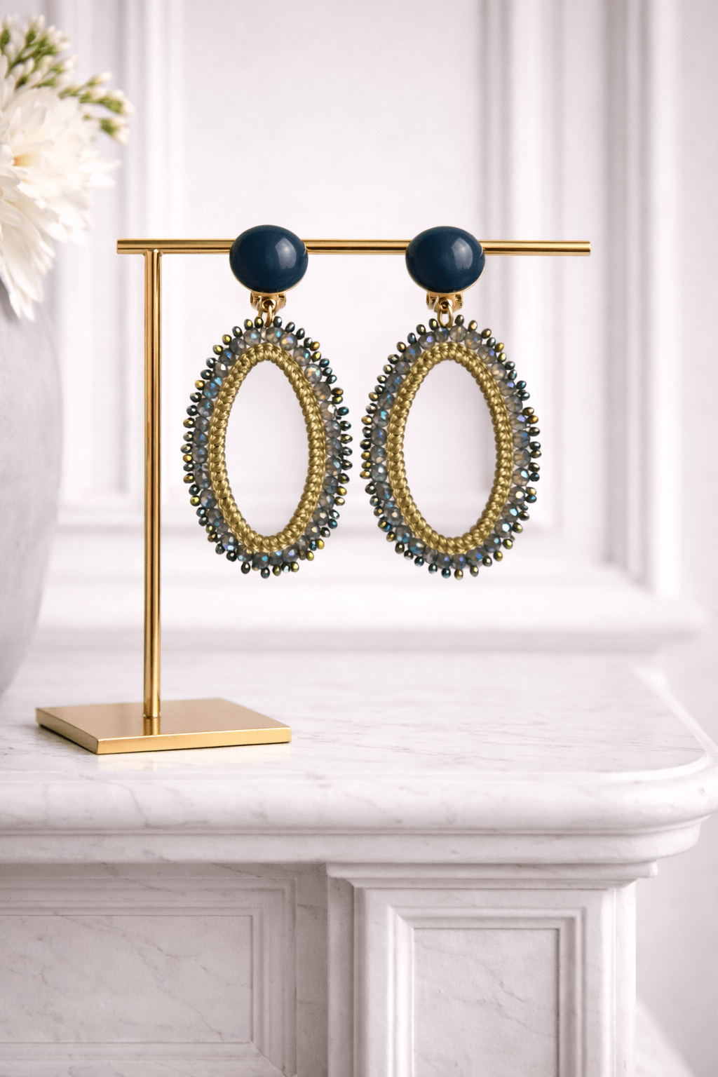 Ovale Clip-Ohrringe mit dunkelblauem Stecker, goldfarbenem Garnrahmen und umlaufenden schimmernden Glasperlen, präsentiert auf einem goldfarbenen Schmuckständer vor hellem Hintergrund.