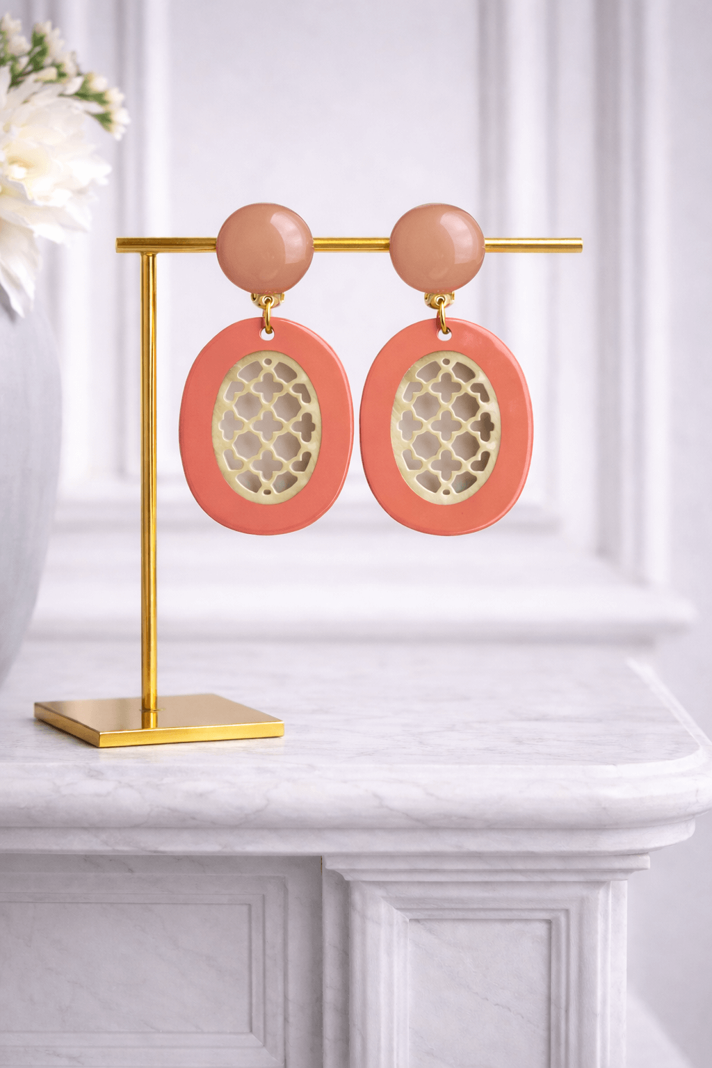 Ovaler Ohrring aus coral lackiertem Büffelhorn mit goldfarbenem ornamentalem Muster und runder Kugel in Nude-Ton auf goldfarbenem Ständer.
